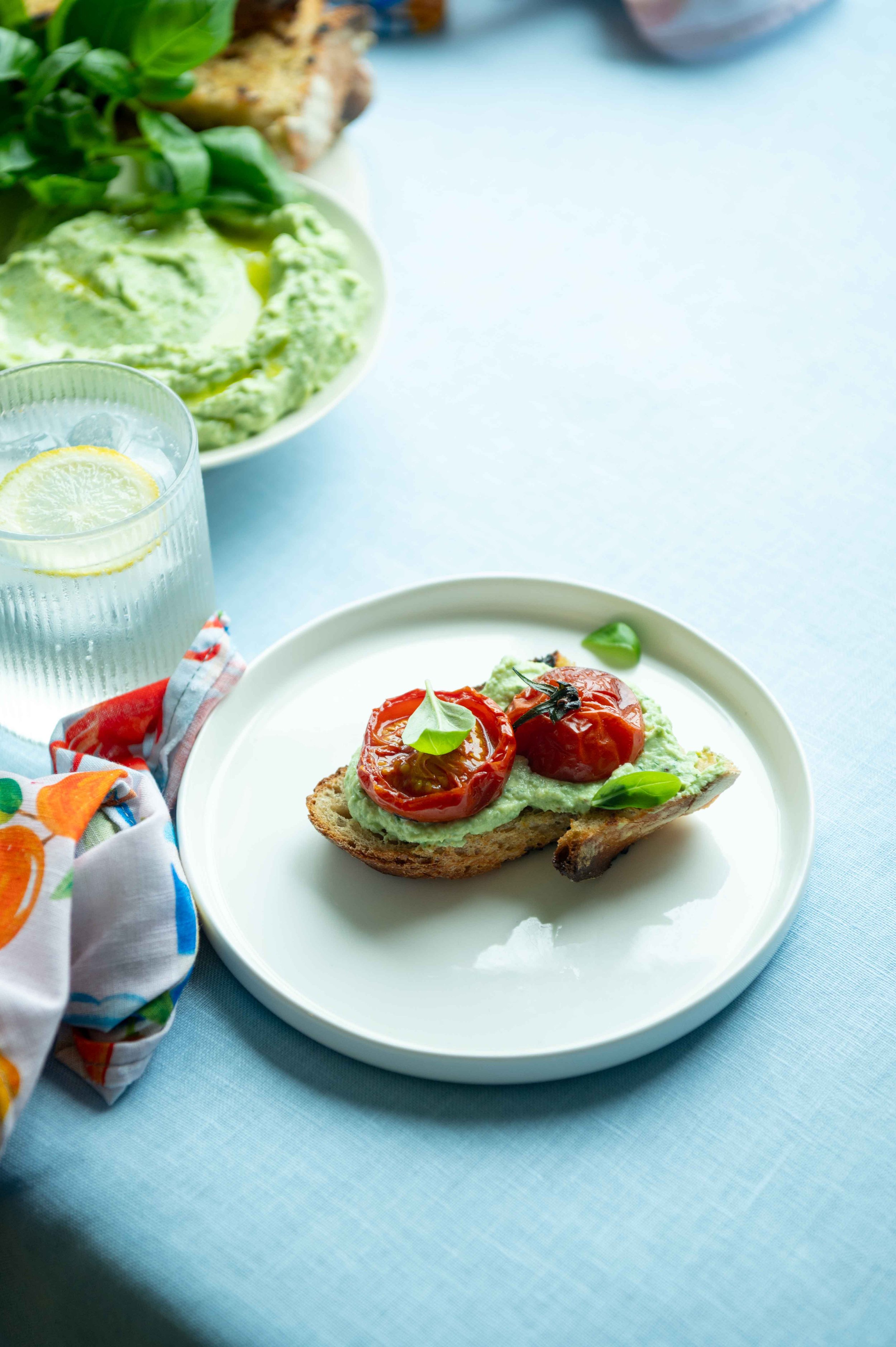 Slow roasted tomatoes on top of green ricotta on sourdough bread. For Life & Leisure Magazine. Shot on a white plate on a table with light-blue linen.