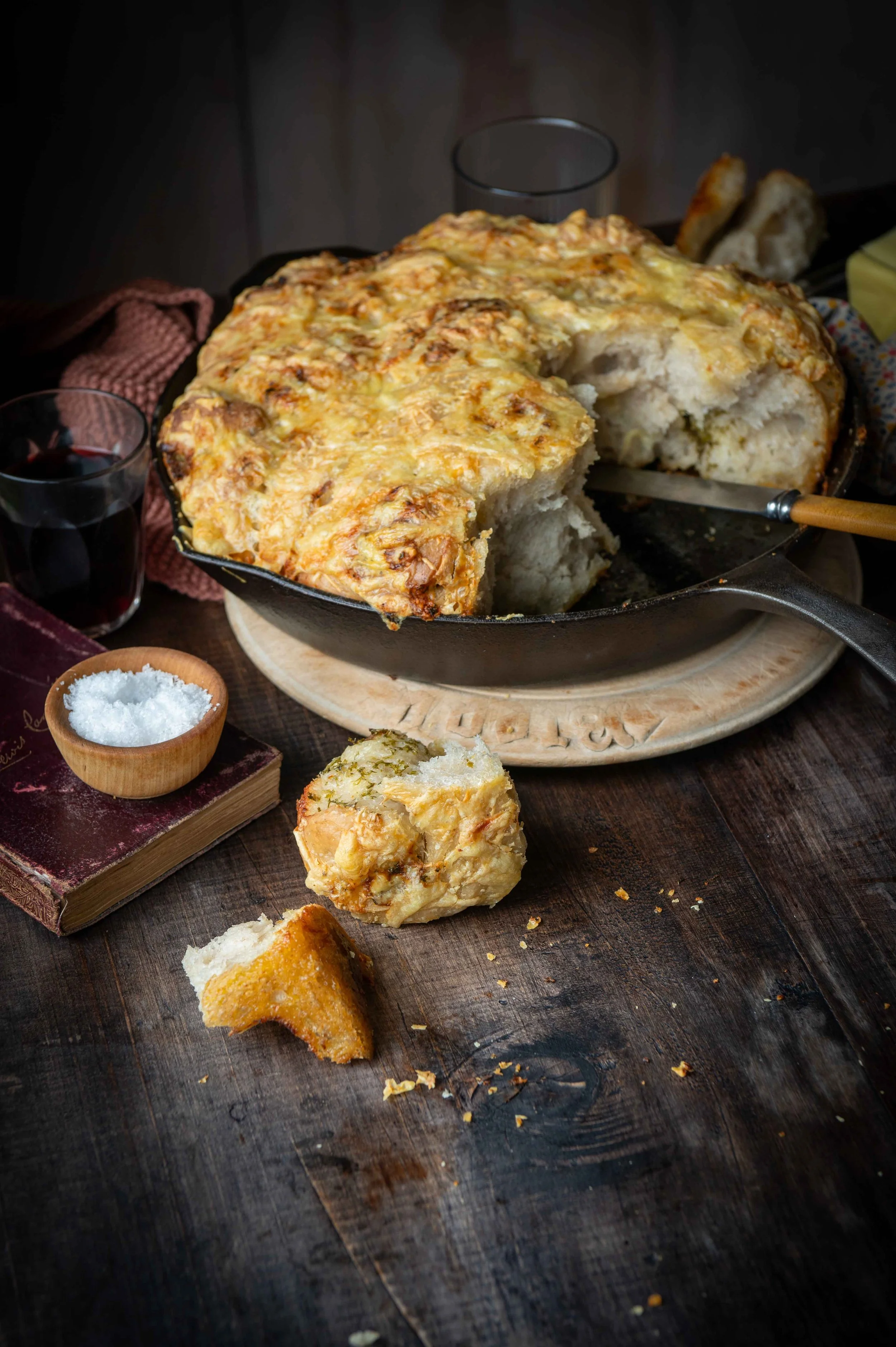 A skillet of freshly baked sourdough garlic pull-apart bread placed on a wooden table along with a small bowl of salt, a glass of red wine, and a pink napkin. The bread has a crispy cheesy golden top. Shot on a dark background.
