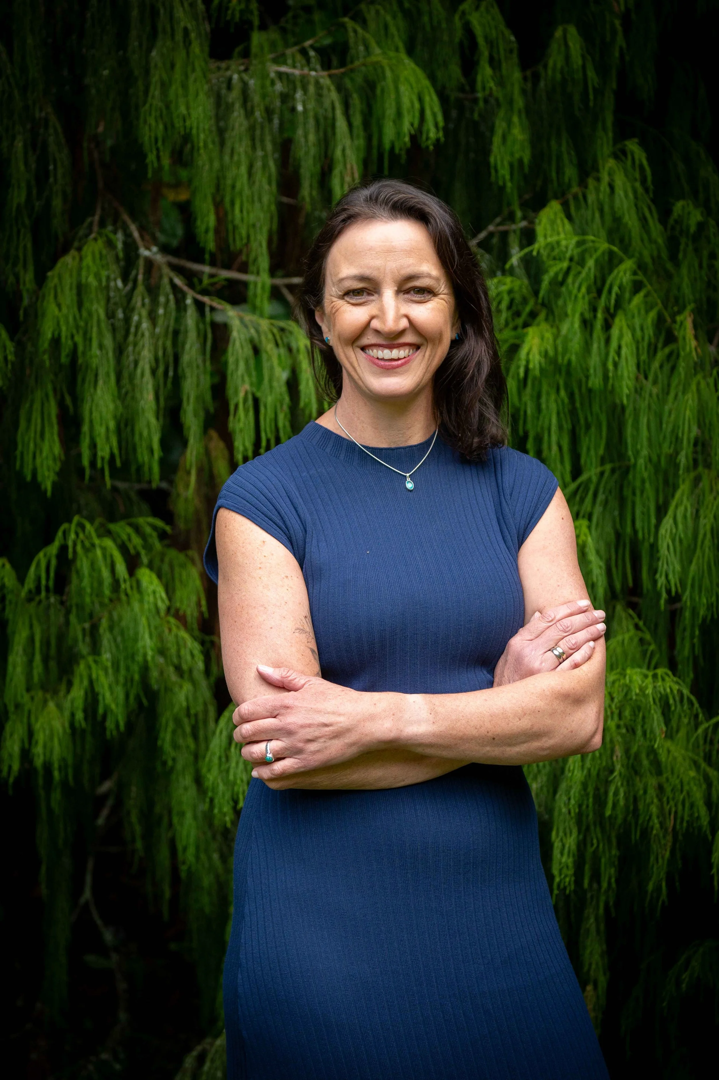A portrait of a woman shot from the waist up against a beautiful green backdrop of rimu trees. She has her arms crossed and is wearing a dark blue knit dress.