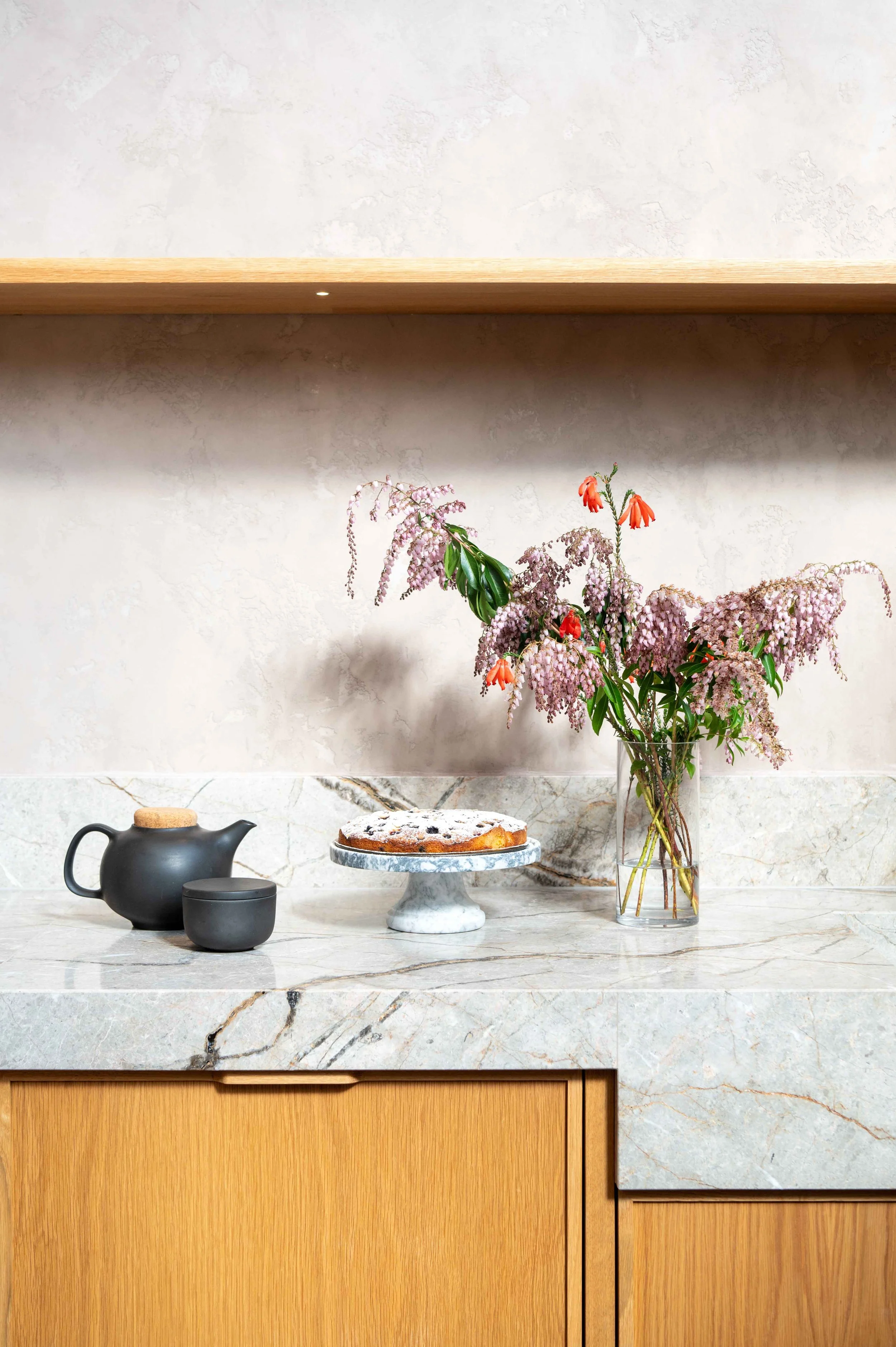 A marble bench top propped with a black teapot, a cake on a cake plate and a vase of pink and orange flowers. Natural light.