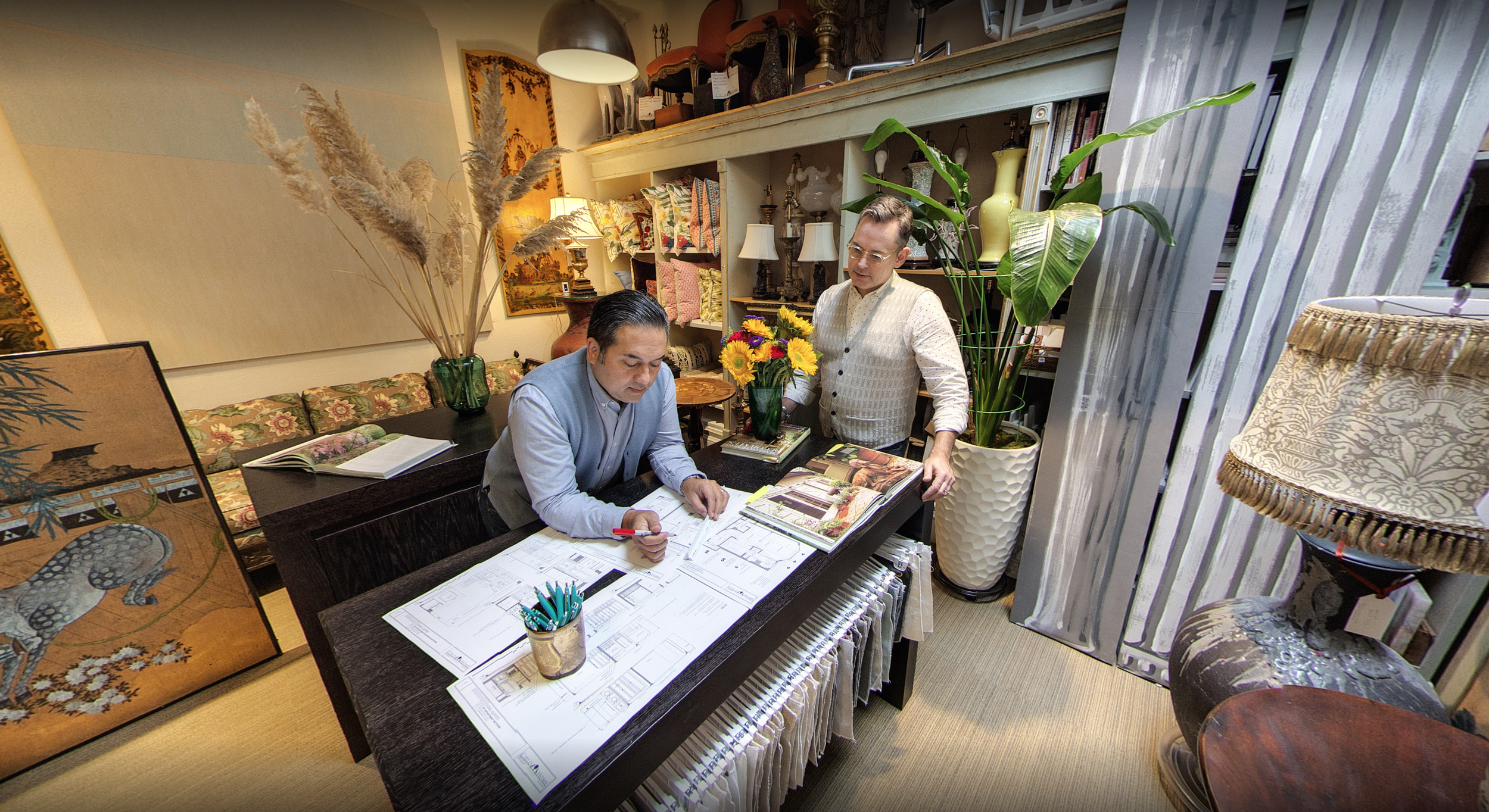 Two men are having a discussion at a table filled with blueprints and design magazines. The room has shelves filled with lamps, vases, and decorative items. One man is seated, reviewing the blueprints with a red pen, while the other stands, holding some magazines. The space is decorated with large plants, lamps, artwork, and a folding screen with a tiger graphic.