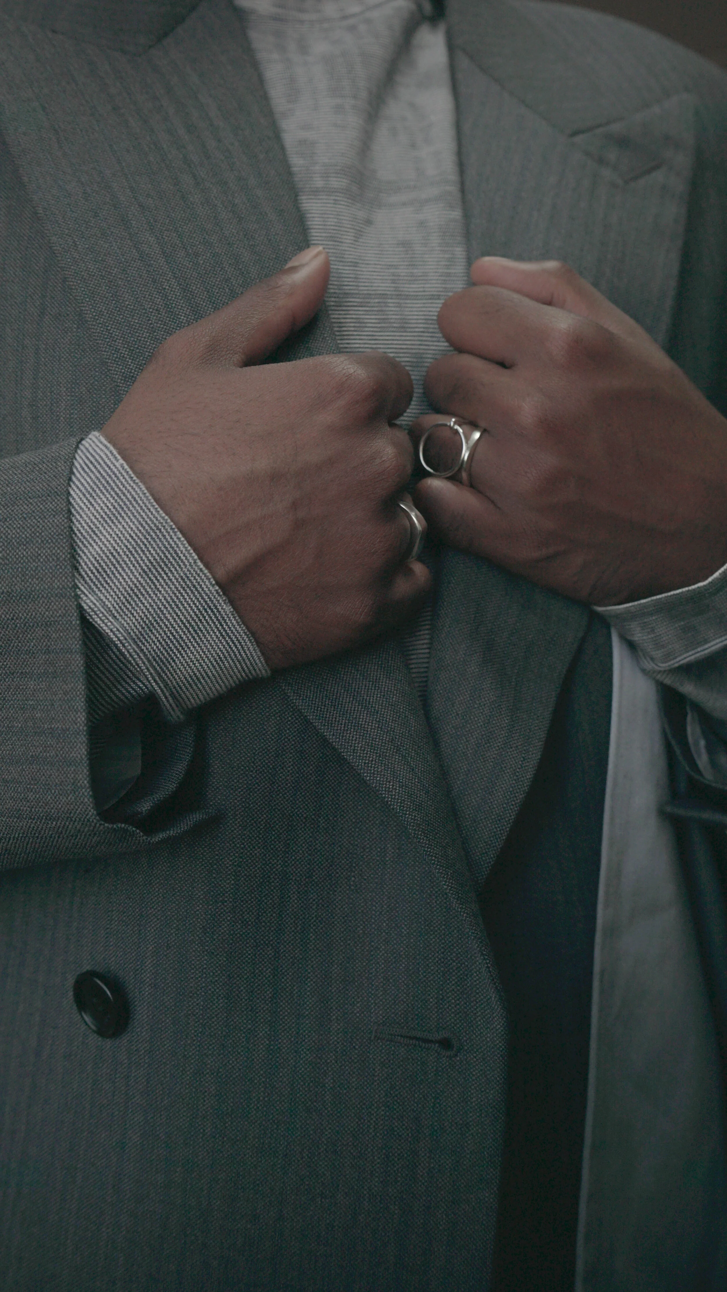 Close-up of a person wearing a gray suit and striped shirt, holding the lapels of the jacket with both hands, wearing rings.