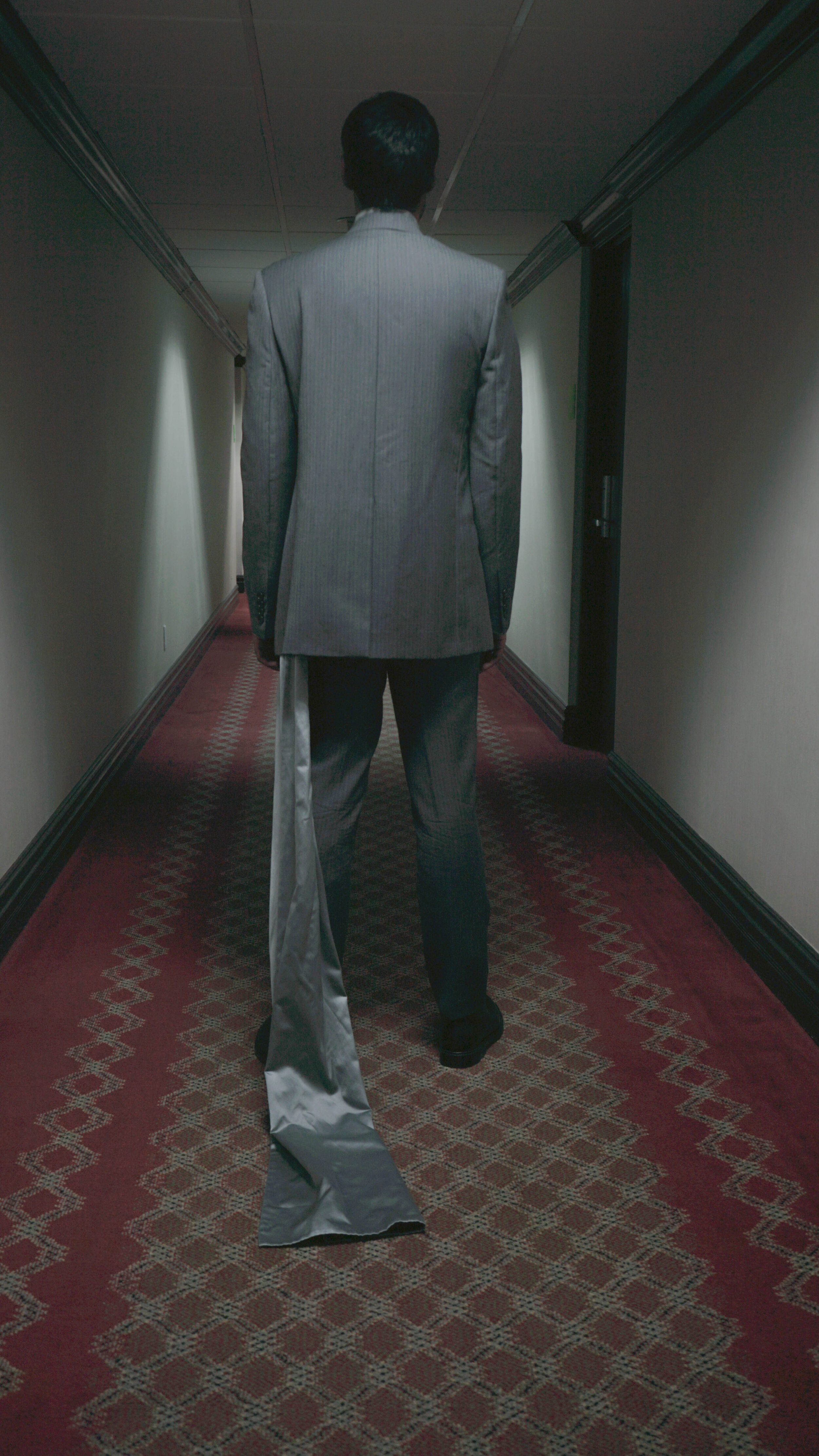 A man in a gray suit walking down a dimly lit hallway with beige walls and a patterned carpet. He has a long, satin-like gray tail attached to his suit.