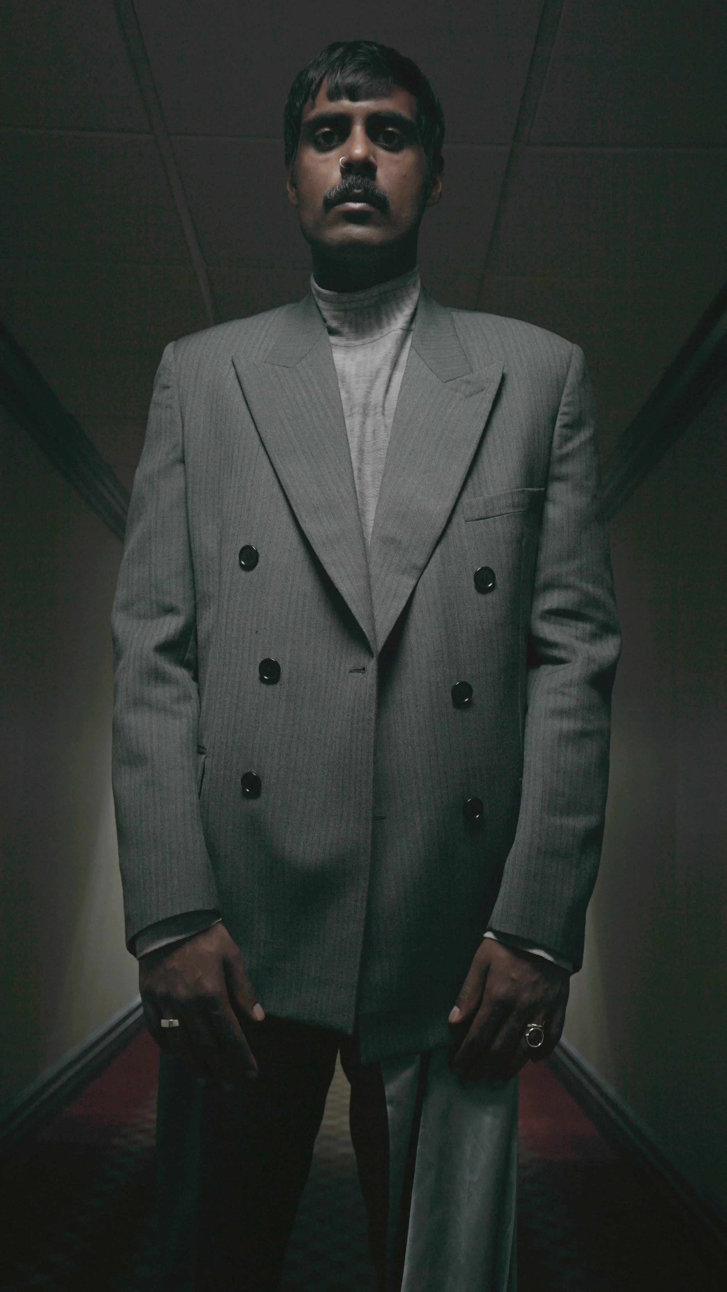 A man with dark hair, mustache, and nose ring wearing a gray pinstripe double-breasted blazer over a turtleneck, standing in a dimly lit hallway.