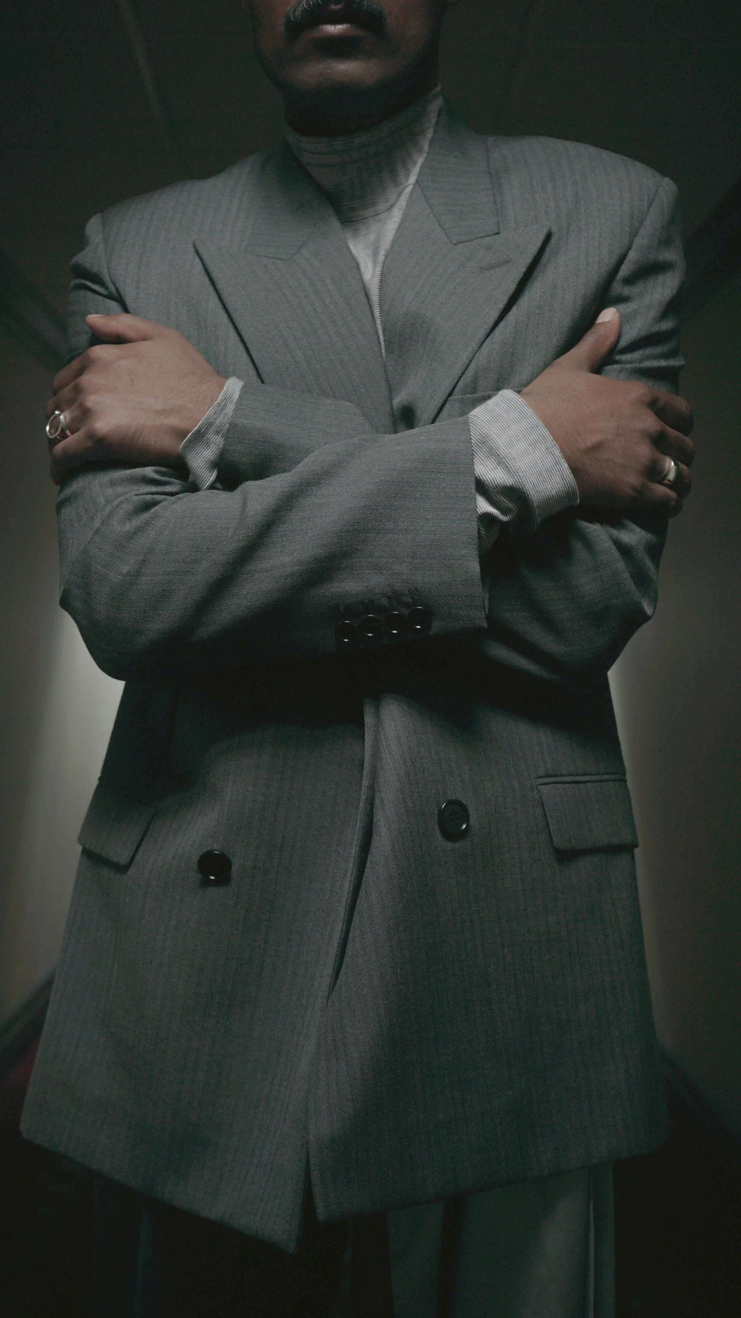 A man in a gray pinstripe suit with crossed arms, standing indoors in a dimly lit environment.