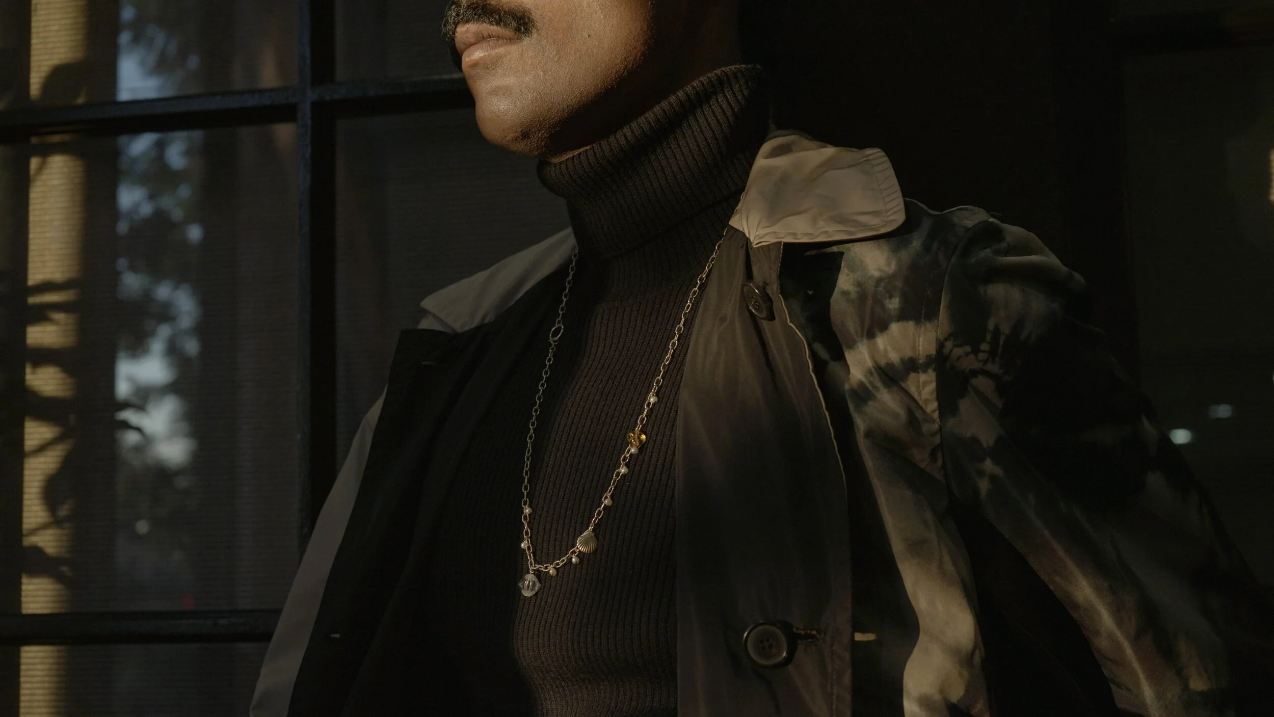 Close-up of a person wearing a black turtleneck, a patterned jacket, and layered necklaces, standing near a window with light reflections.