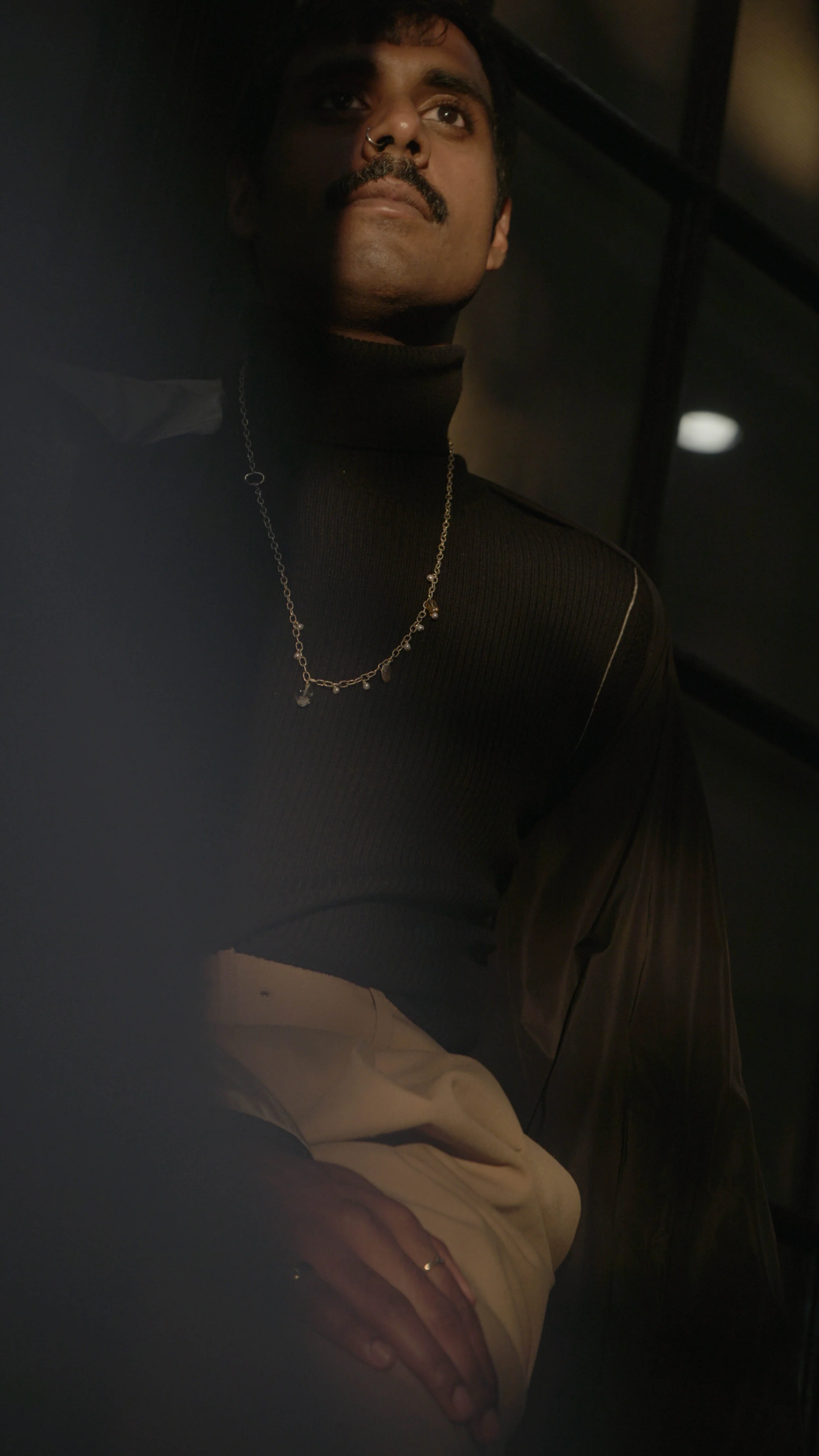 A man with dark hair, a mustache, and a nose piercing wearing a black turtleneck and silver chain necklace seen in a dimly lit setting.