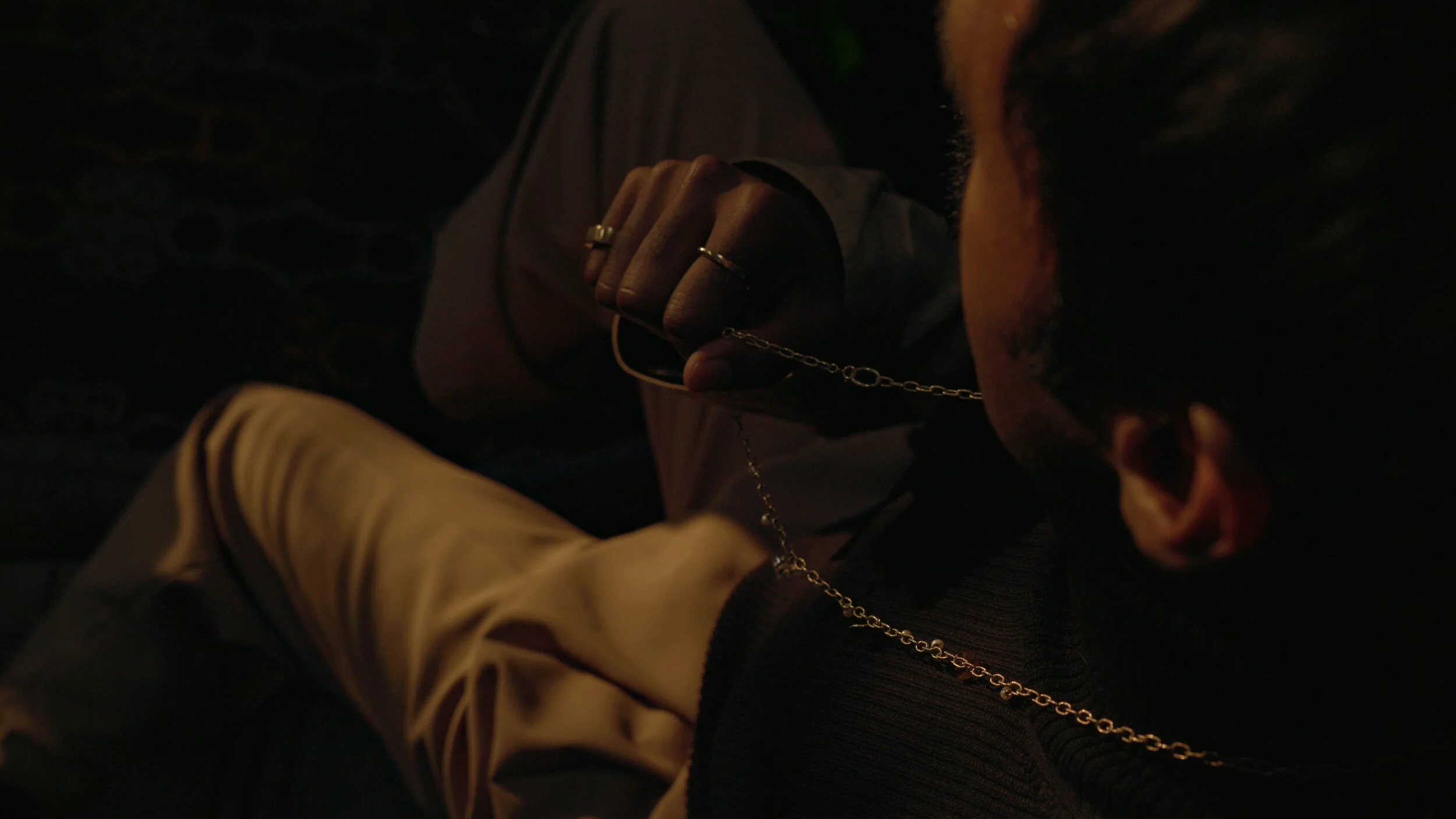 Person wearing a Dries Van Norten Coat and black shirt, holding a Ulsi Omenchain necklace, with rings on fingers, in a moody setting