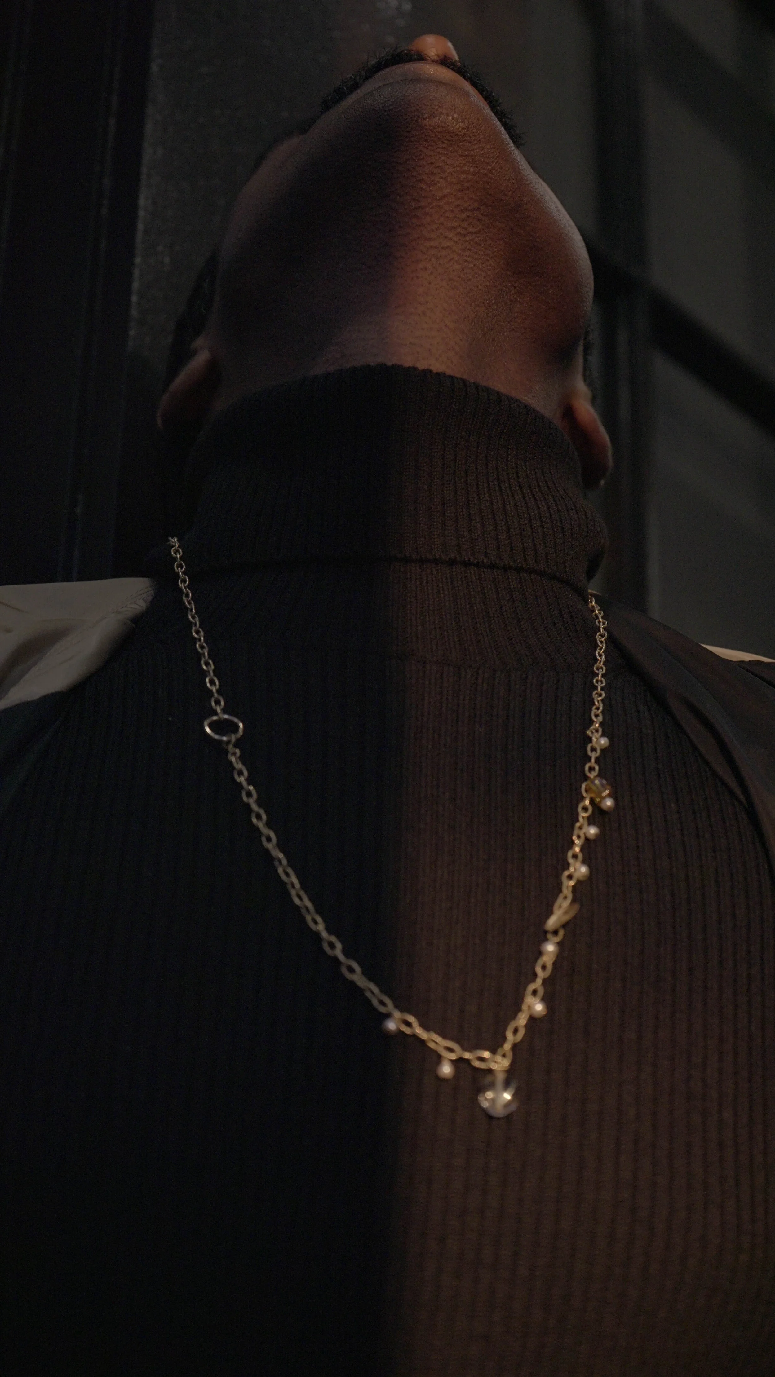 Close-up of a person wearing a black turtleneck and an ulsi omen gold necklace with pearls, shown from below and behind.