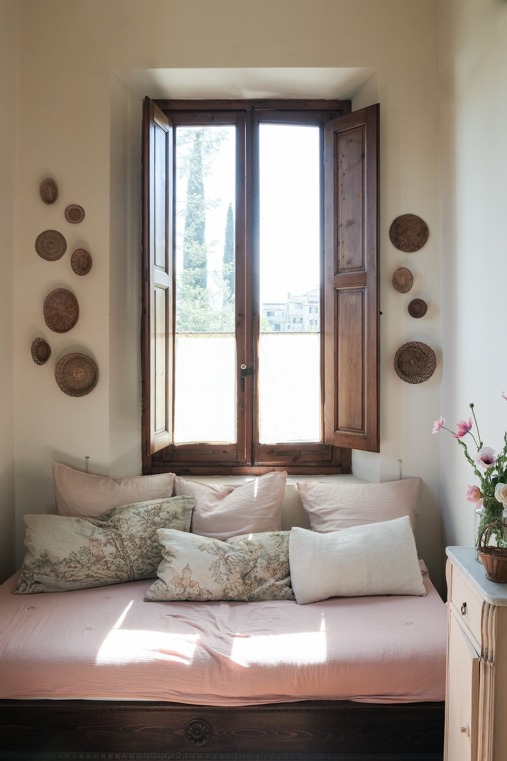 Interno di una stanza con una grande finestra a doppia anta in legno, decorata con piatti e cuscini su un divano, e una piccola lampada a fianco.