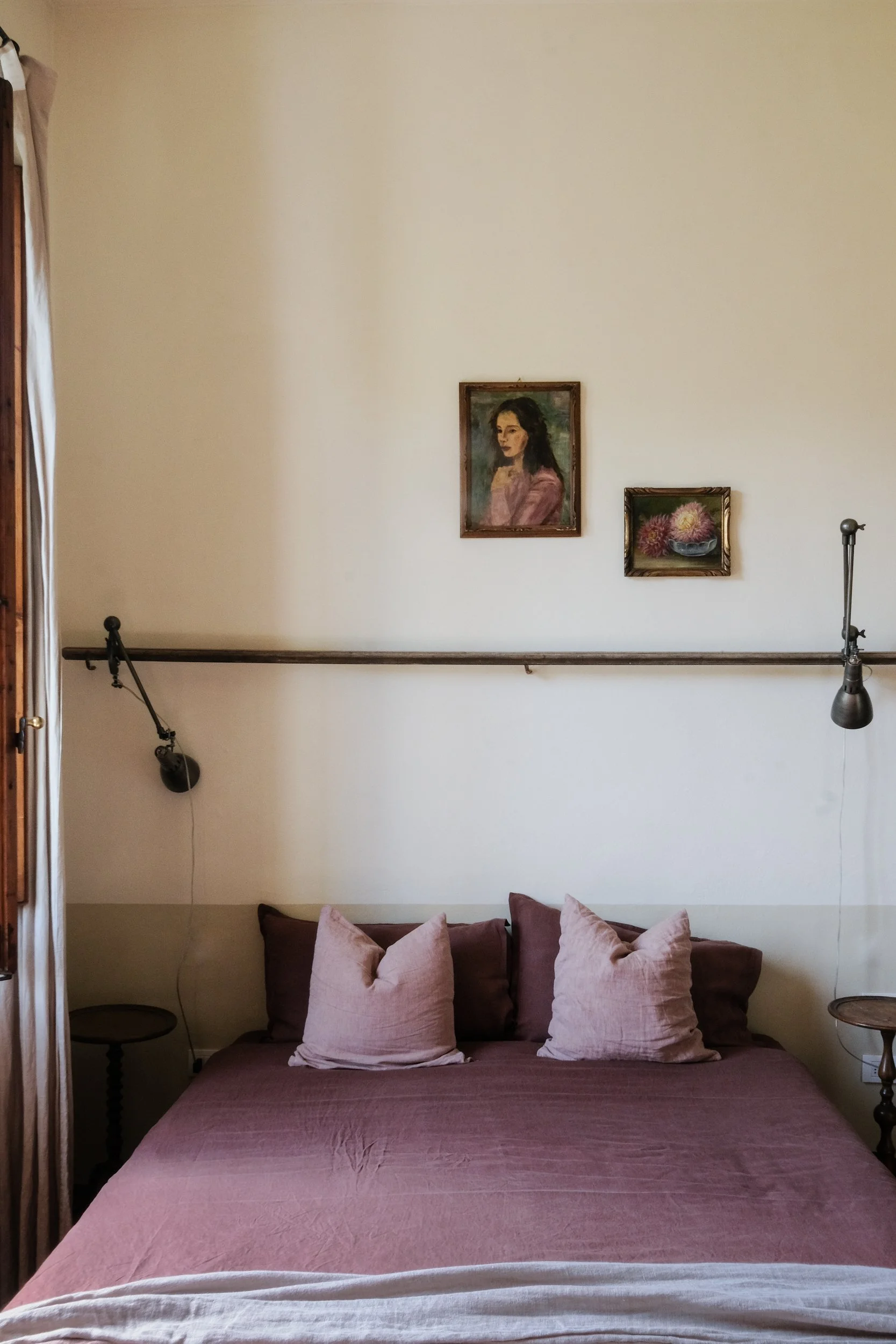 Una stanza con un letto matrimoniale con copriletto rosa e cuscini rosa chiaro. Sopra il letto ci sono due quadri appesi su una parete beige: uno ritrae un ritratto di donna e l'altro un vaso di fiori. Alla sinistra ci sono tende con drappeggi e un'illuminazione da lampada da parete.