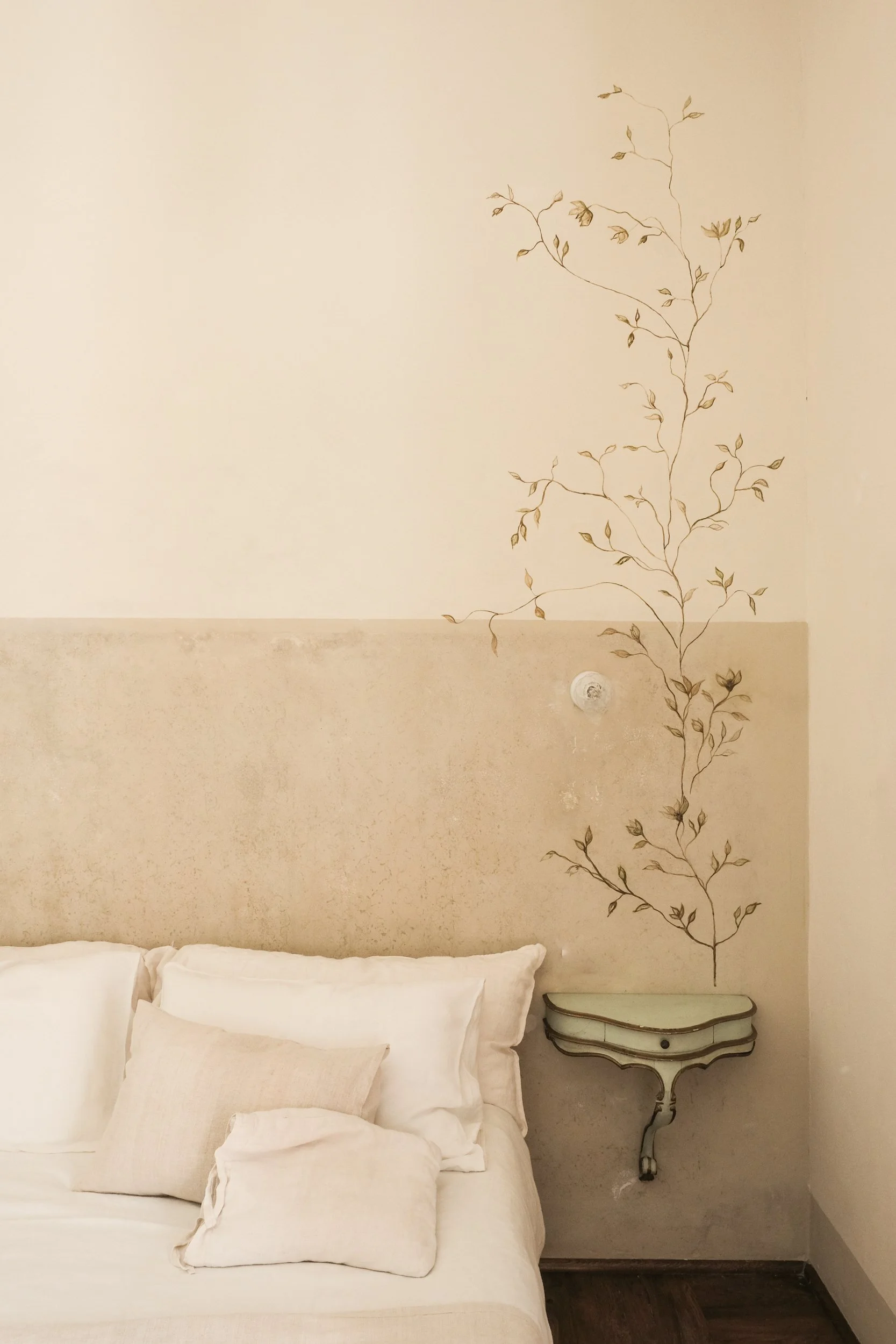 Letto matrimoniale con cuscini bianchi, fianco di una parete decorata con un disegno di un ramoscello e foglie, e una piccola consolle di colore chiaro con disegni stilizzati.