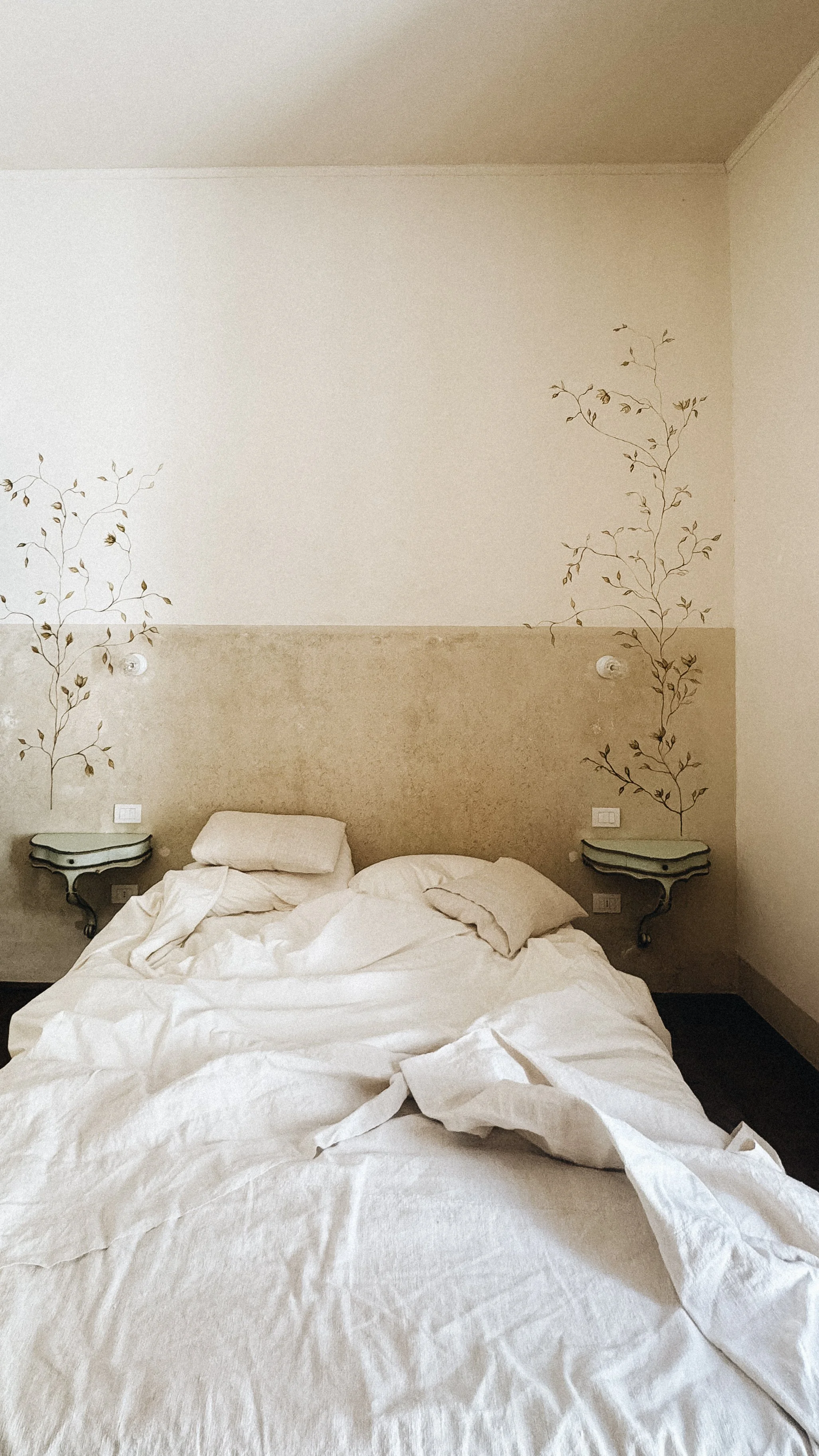 Letto disordinato con cuscini e lenzuola sbagliate, due lampade da parete e due piccole mensole nere accanto al letto, decorazioni di rami con foglie sul muro.