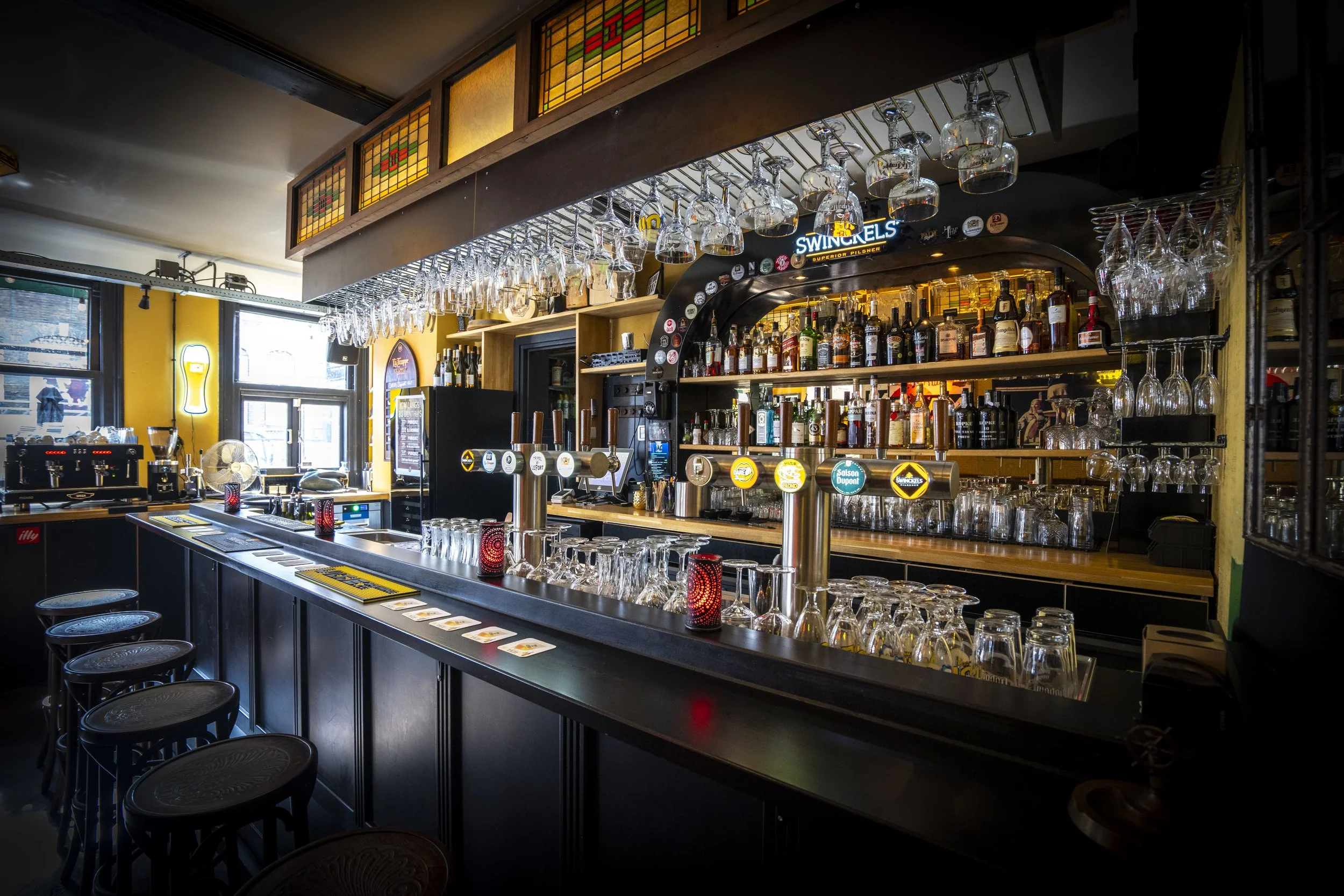 Interieur van een gezellige bar met een donker houten barcounter, rum- en wodkaflessen op de achtergrond, glaswerk boven de bar en enkele barkrukken voor de counter.