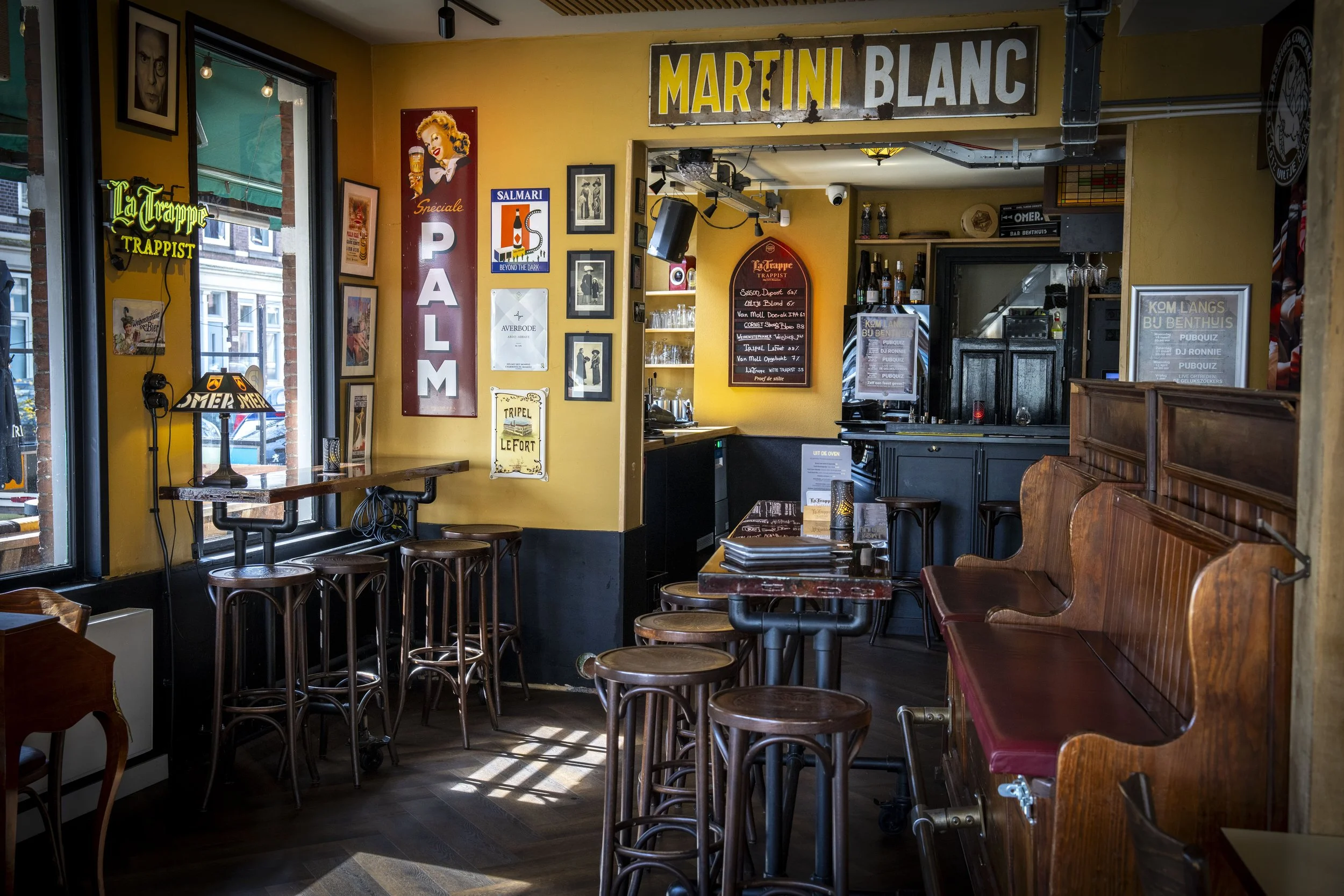 Interieur van een café met houten banken, hoge krukken, muurschilderingen en posters, waaronder een grote gele 'MARTINI BLANC' banner.