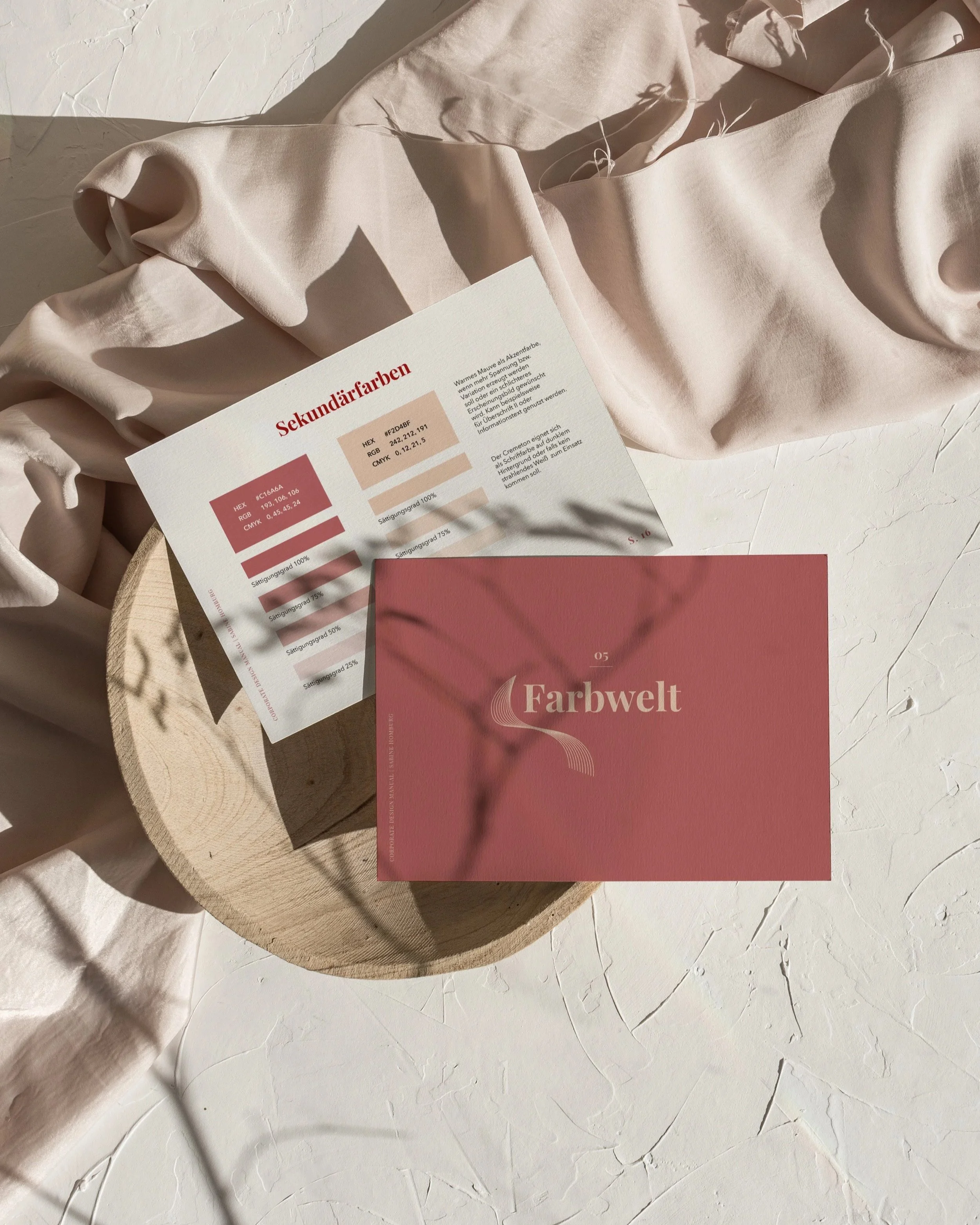 Eine digitale Farbkarte mit dem Titel 'Sekundärfarben' und einem Farbton namens 'Farbwelt', die auf einem runden Holztablett liegt, umgeben von einem beige Stoff und Schatten auf einer weißen, strukturierten Oberfläche.