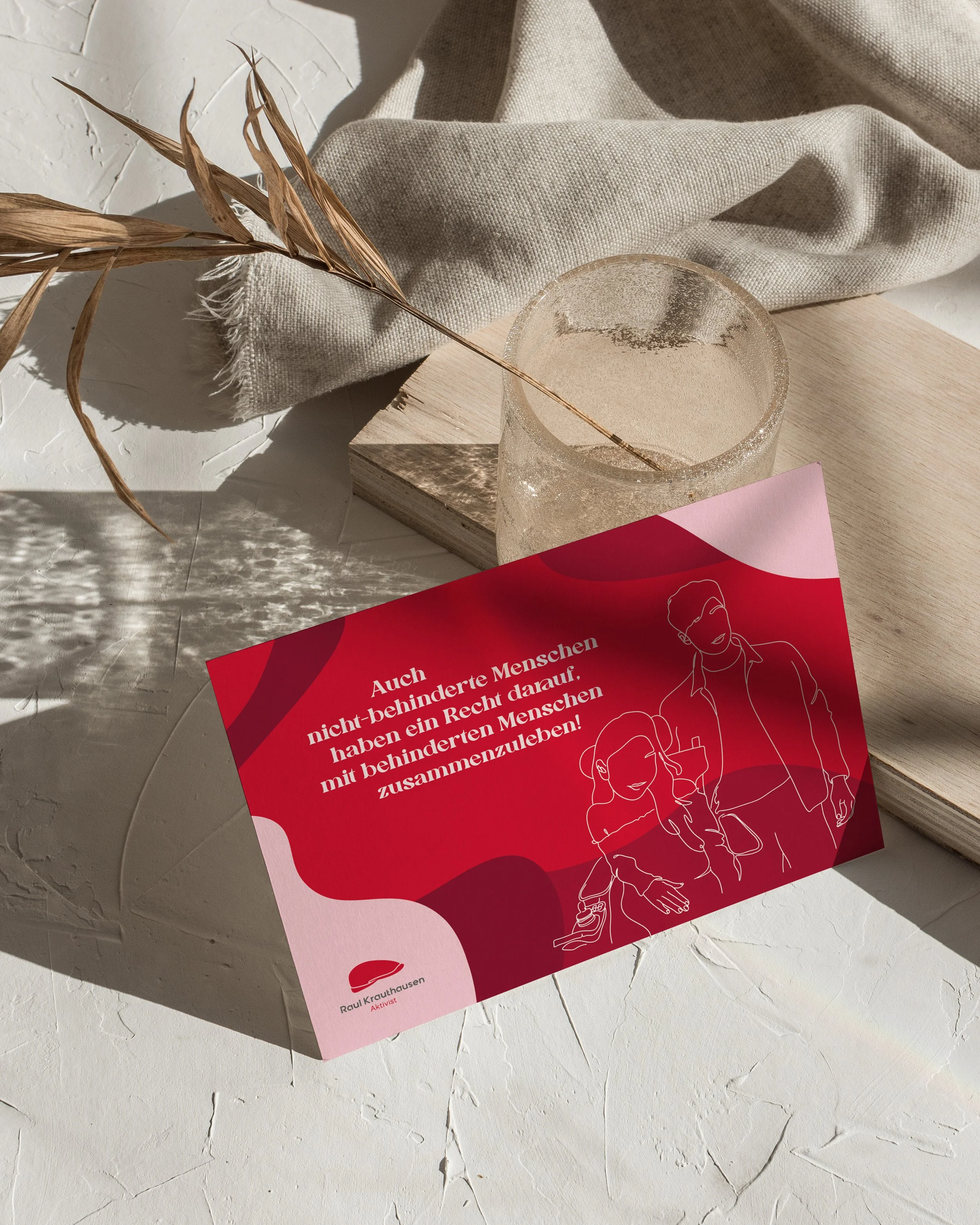 Ein rotes Plakat mit weißen Texten und einer einfachen Linienzeichnung einer behinderten Person, die von einer Begleitperson unterstützt wird, liegt auf einer weißen Oberfläche neben einem leeren Glas und einer Stoffbahn.