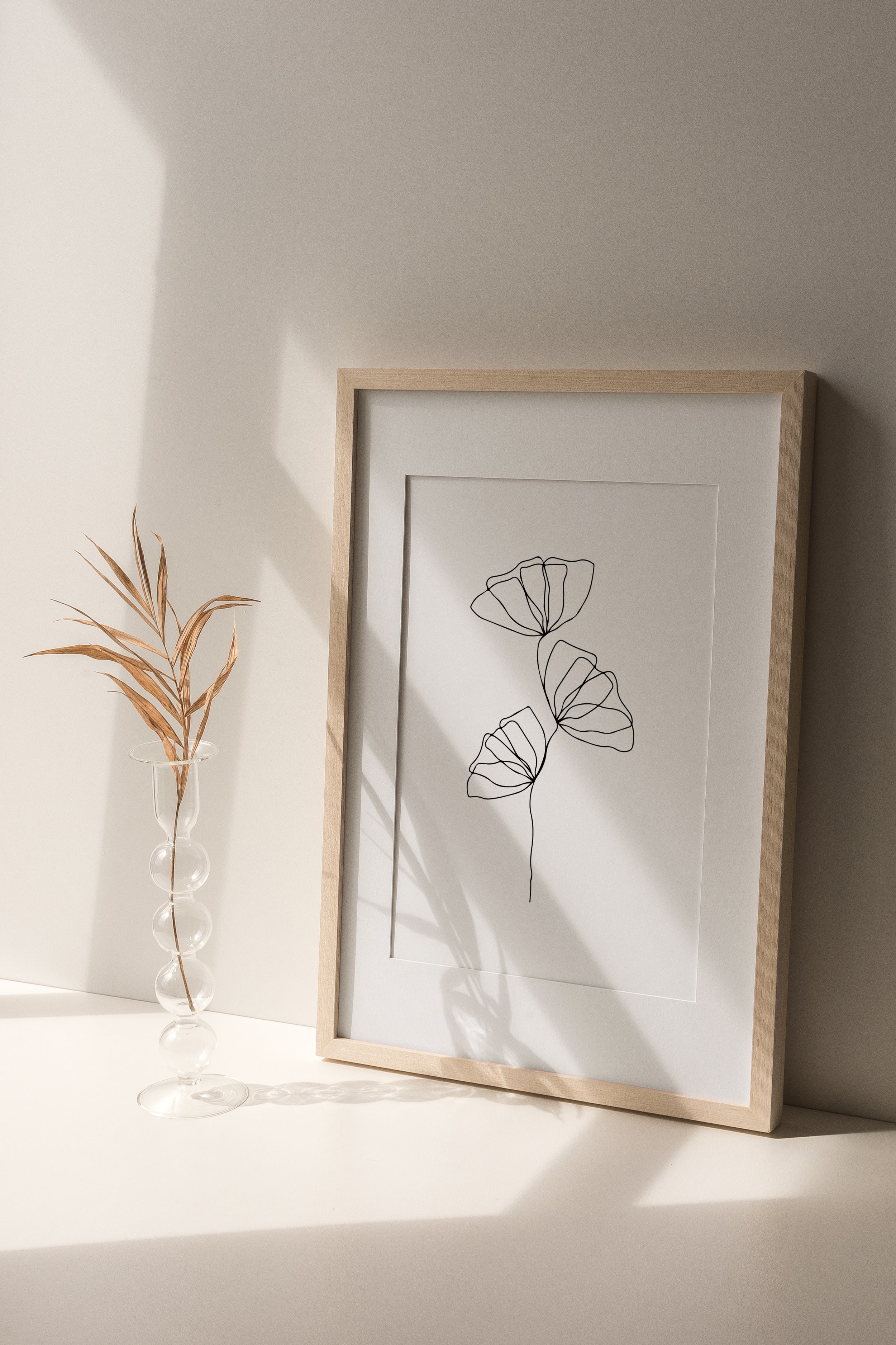 Gerahmte minimalistische Linienzeichnung einer Pflanze auf weißem Hintergrund, neben einer Glasvase mit getrockneten Blättern im sanften Tageslicht.