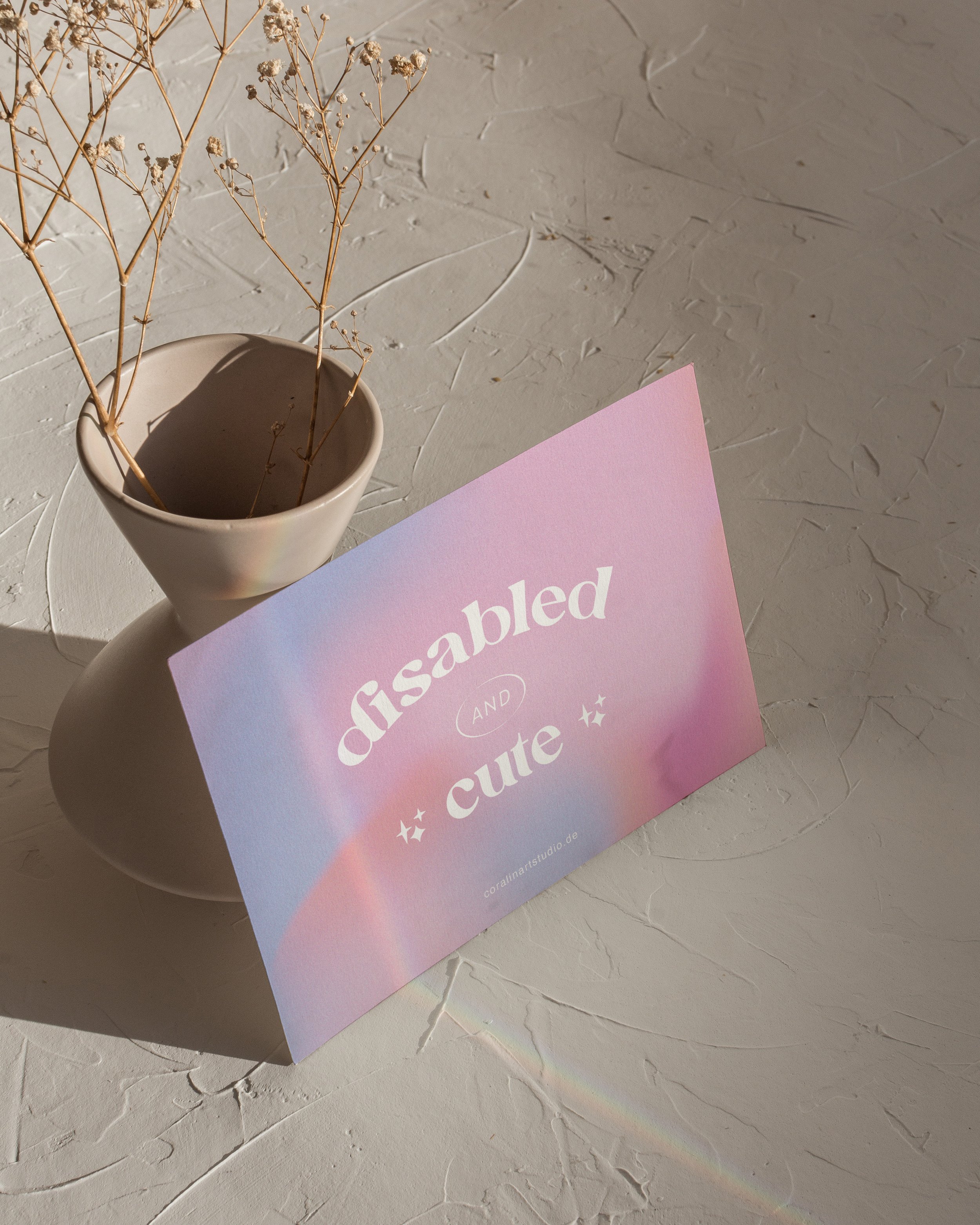 Postkarte mit pastellfarbenem Verlauf und dem Text 'disabled AND cute' neben einem weißen Blumentopf mit getrockneten Zweigen auf strukturiertem Untergrund
