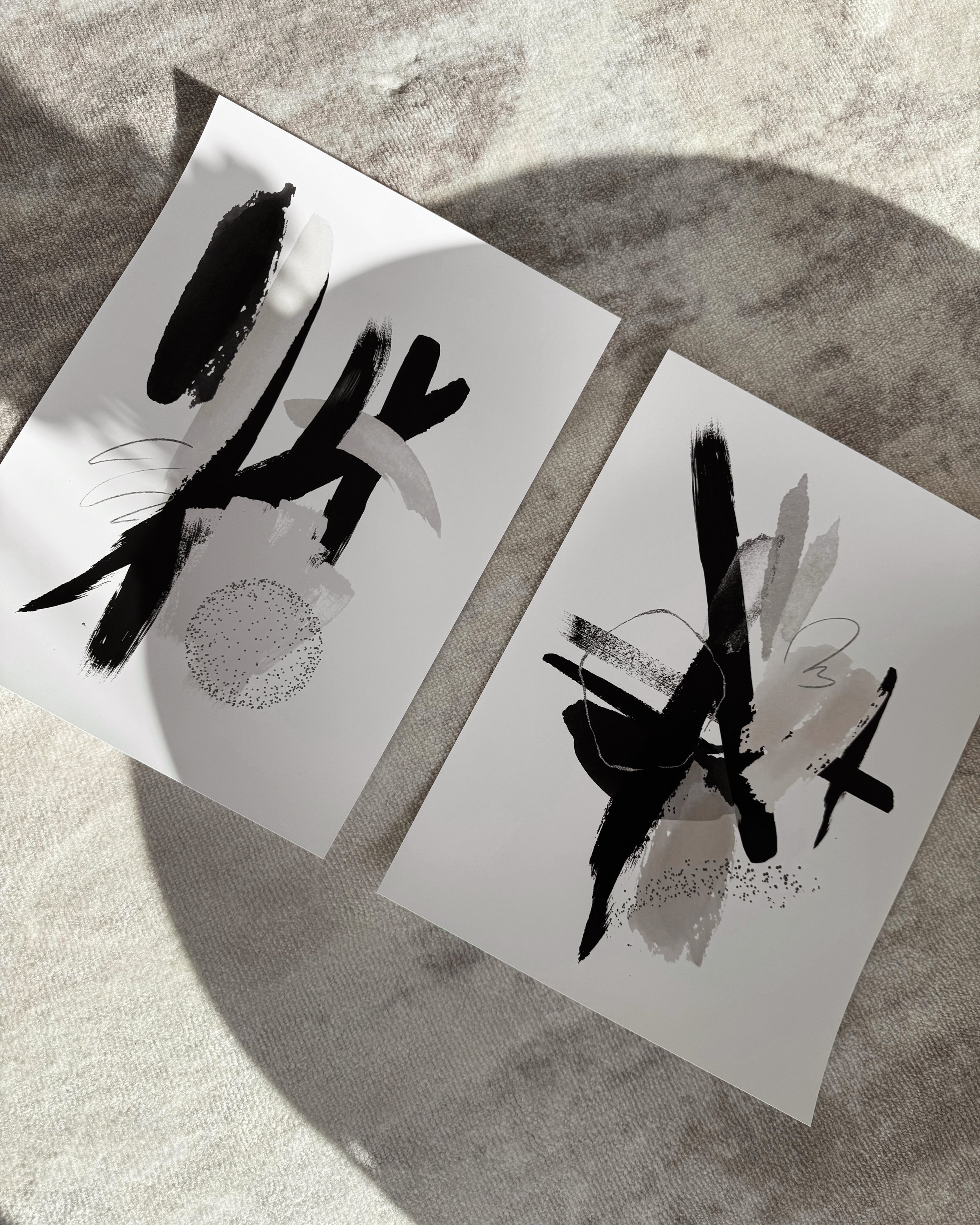 Zwei Prints mit abstrakten, schwarz-weißen Linienzeichnungen liegen nebeneinander auf einem hellen, strukturierten Untergrund.