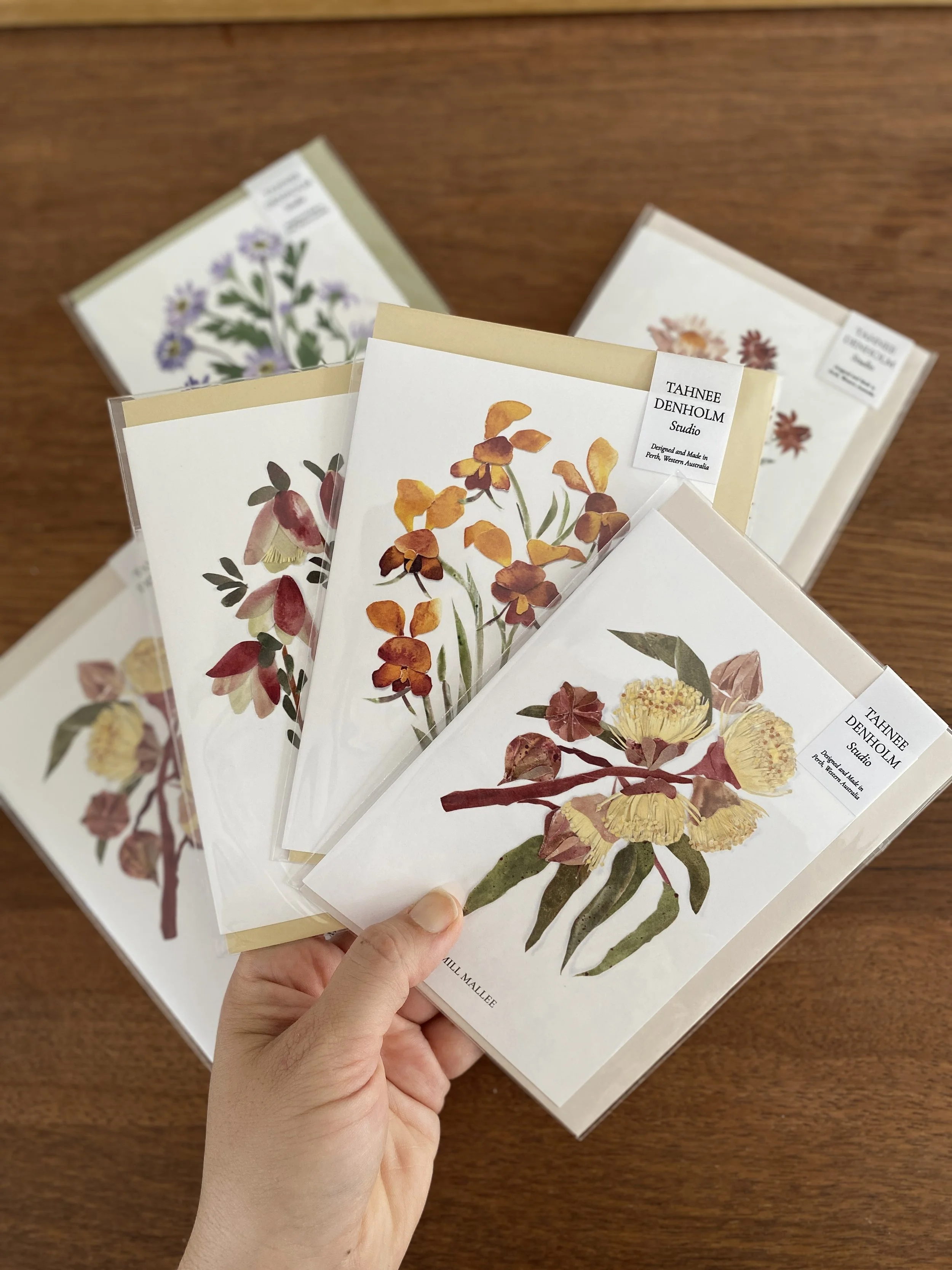 Hand holding five botanical illustration cards with floral paintings, titled by artist Tahnee Denholm, with three additional floral illustration cards in the background on a wooden surface.