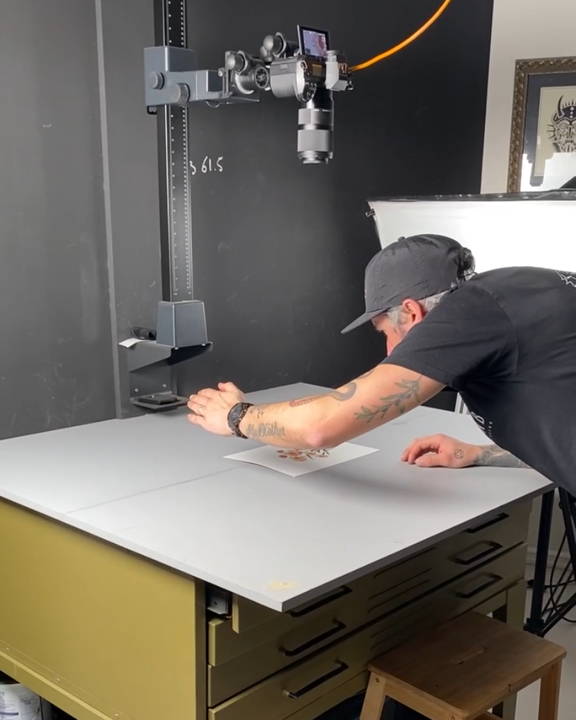 A person with tattoos, wearing a black cap and t-shirt, is doing a photoshoot with a camera mounted on a robotic arm, in a studio setting. The person is leaning over a table with printed artwork.