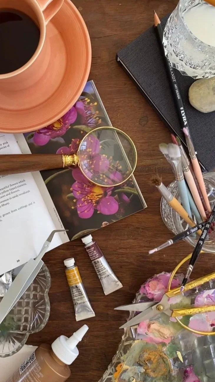 An artist's workspace with a cup of black coffee, a magnifying glass, a notebook, paint tubes, scissors, a paint palette, a book with a flower photograph, and various brushes on a wooden table.