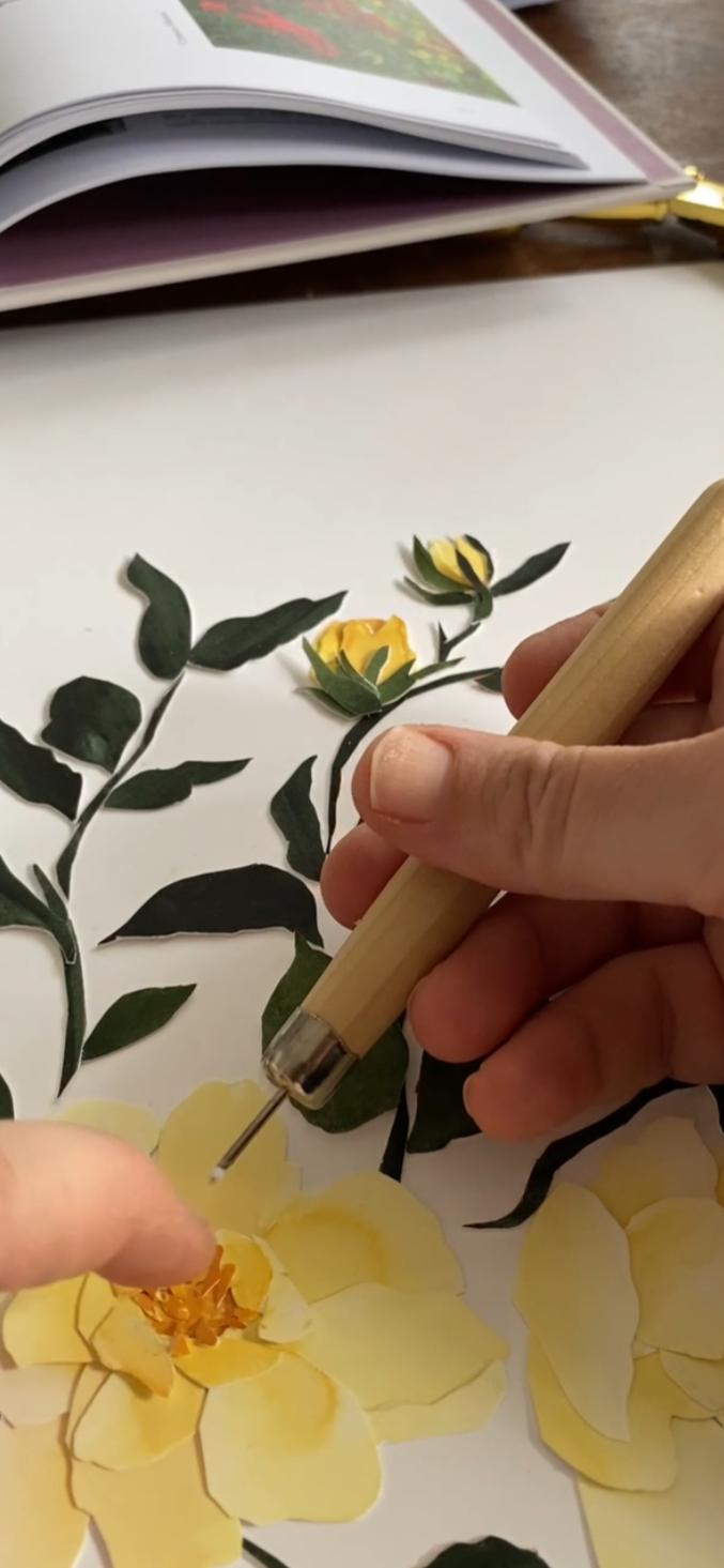 Person using a craft tool on a yellow paper flower, surrounded by paper greenery and an open book on a white surface.