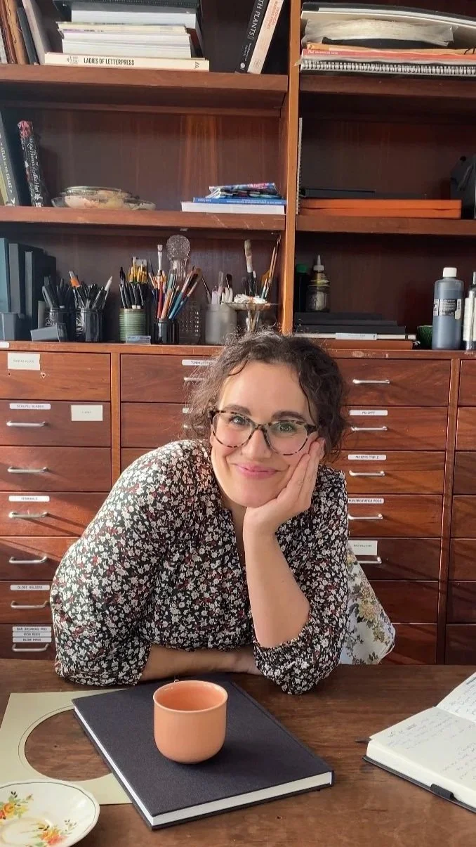 A woman with glasses and curly hair sitting at a wooden desk, resting her chin on her hand, with books, notebooks, and art supplies on shelves behind her.