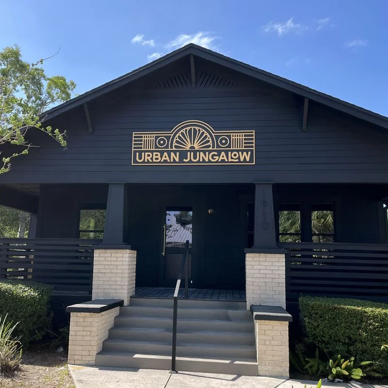 A dark blue building with a sign that reads "Urban Jungalow" and a decorative graphic of a sunburst. The building has steps leading up to a porch with a handrail, and is surrounded by greenery under a blue sky.
