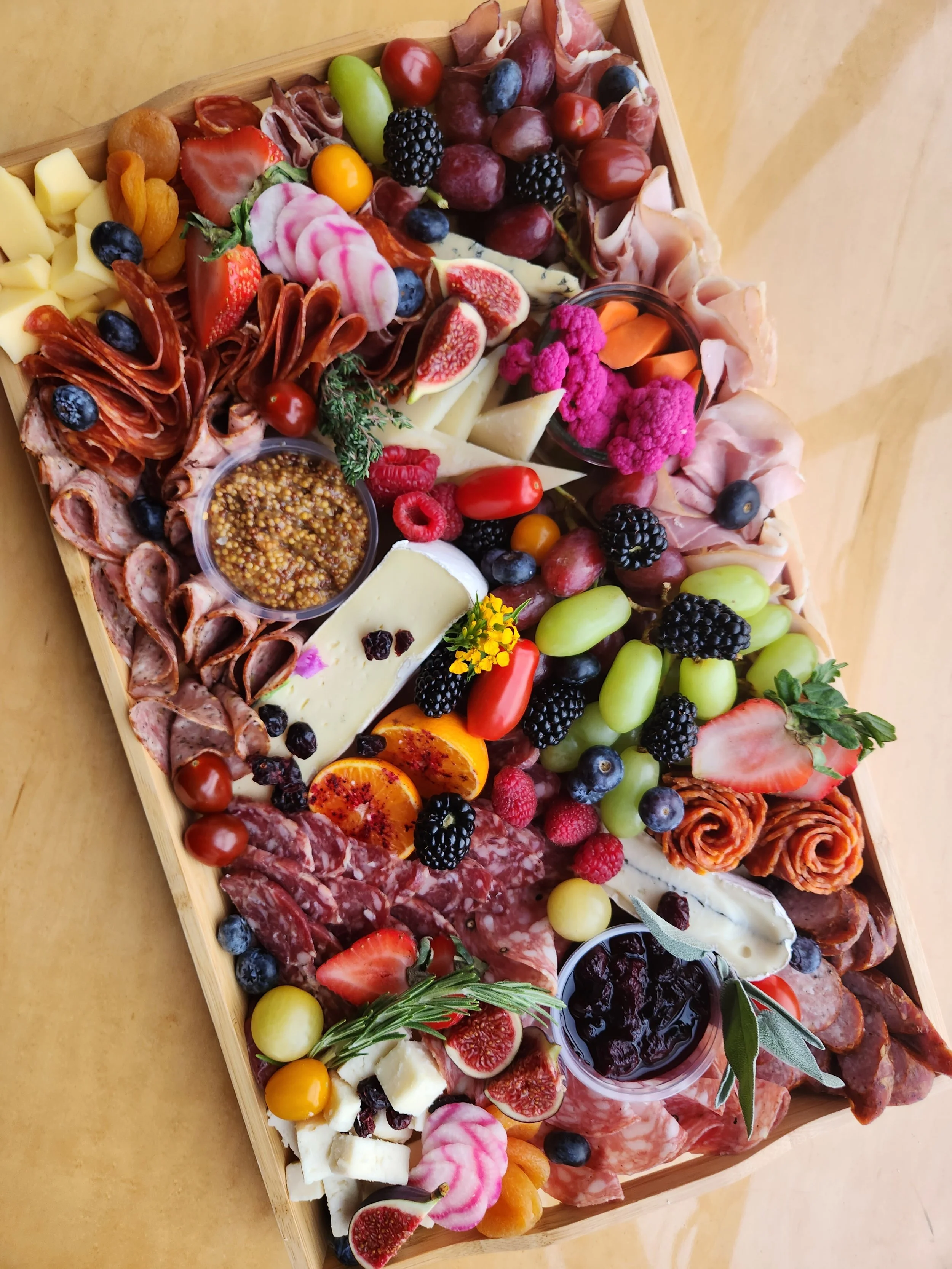 A large assortment of cheeses, fresh fruits like strawberries, blueberries, blackberries, grapes, figs, and sliced oranges, as well as cured meats such as salami, ham, and prosciutto, all arranged on a wooden serving board for a charcuterie platter.