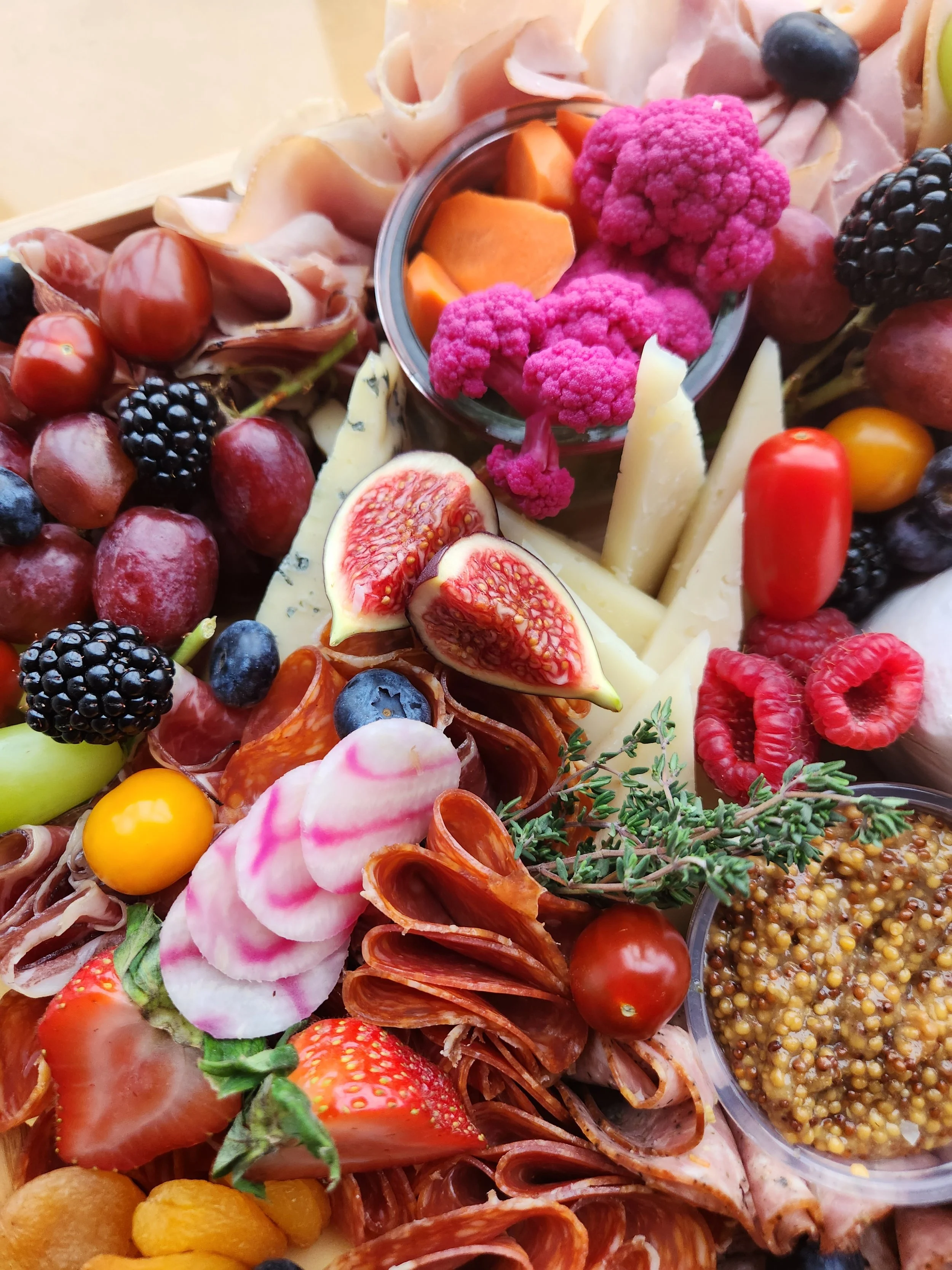 An assorted platter of fresh fruits, cheeses, sliced meats, and a small bowl of mustard seeds.