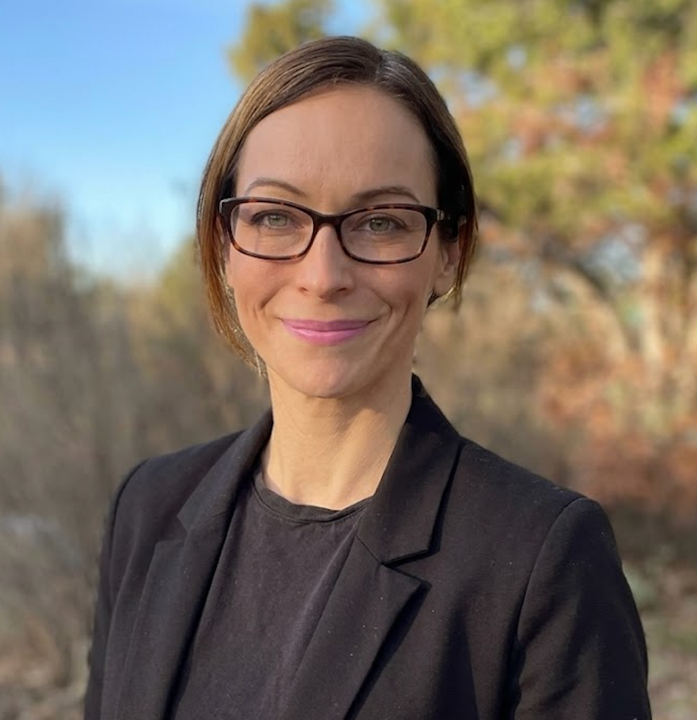 A woman brown hair, glasses, and a black blazer smiling outdoors in a park with trees in the background.
