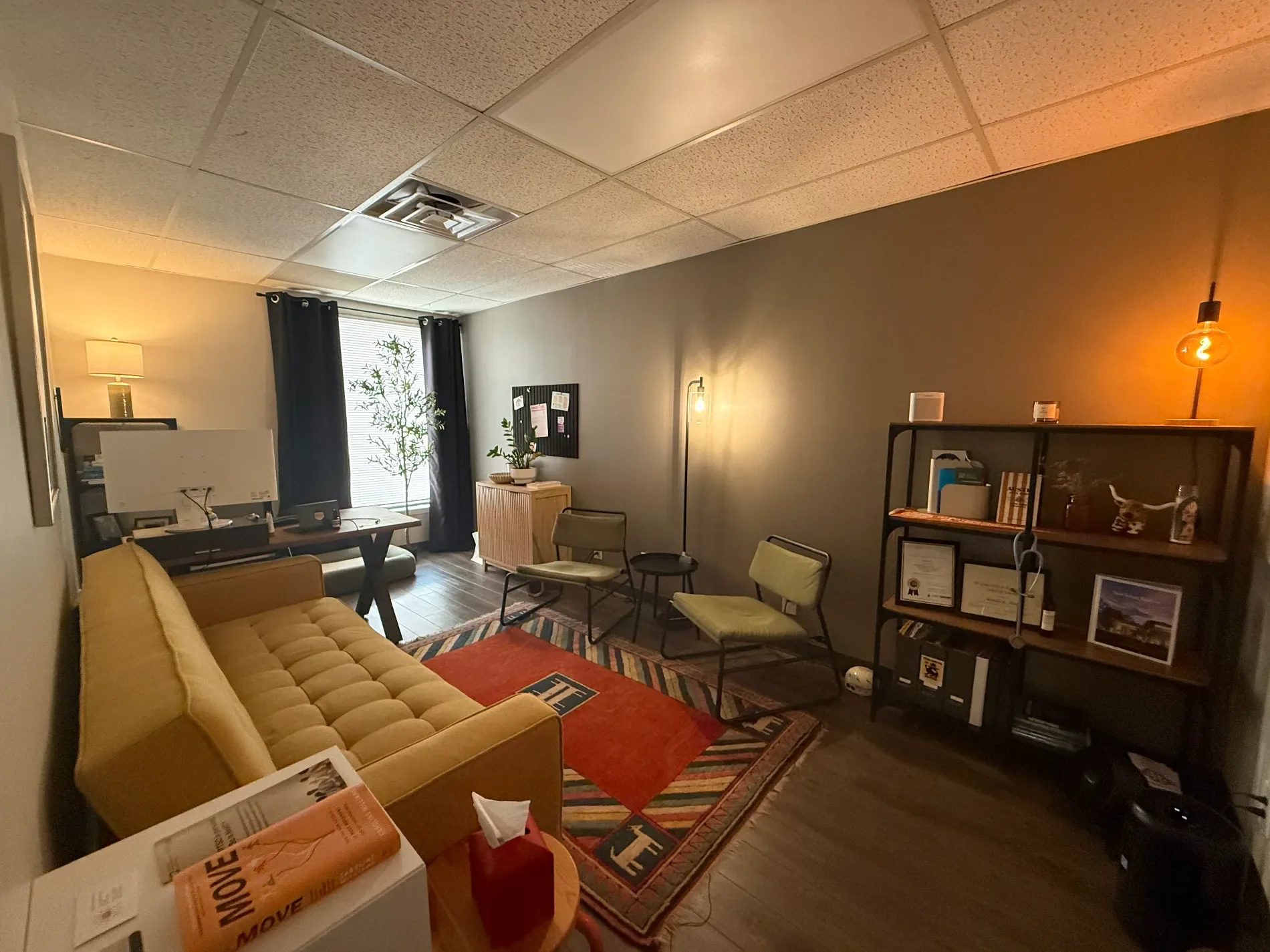 Office or therapy room with yellow tufted sofa, several chairs, a black bookshelf with framed certificates, plants, and a rug with red and black patterns. Warm lighting from a ceiling bulb and a standing lamp. There is a window with dark curtains.