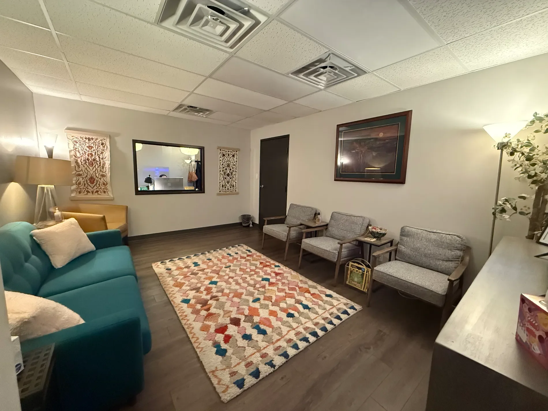 A cozy living room with a teal sofa, beige armchair, and three gray chairs arranged along the wall. There is a colorful patterned rug on the dark wood floor. The walls are decorated with two framed pictures and a decorative fabric wall hanging. A window with a view into an office space is visible. The ceiling has white acoustic tiles with ventilation grilles, and there are various lamps and plants in the room.