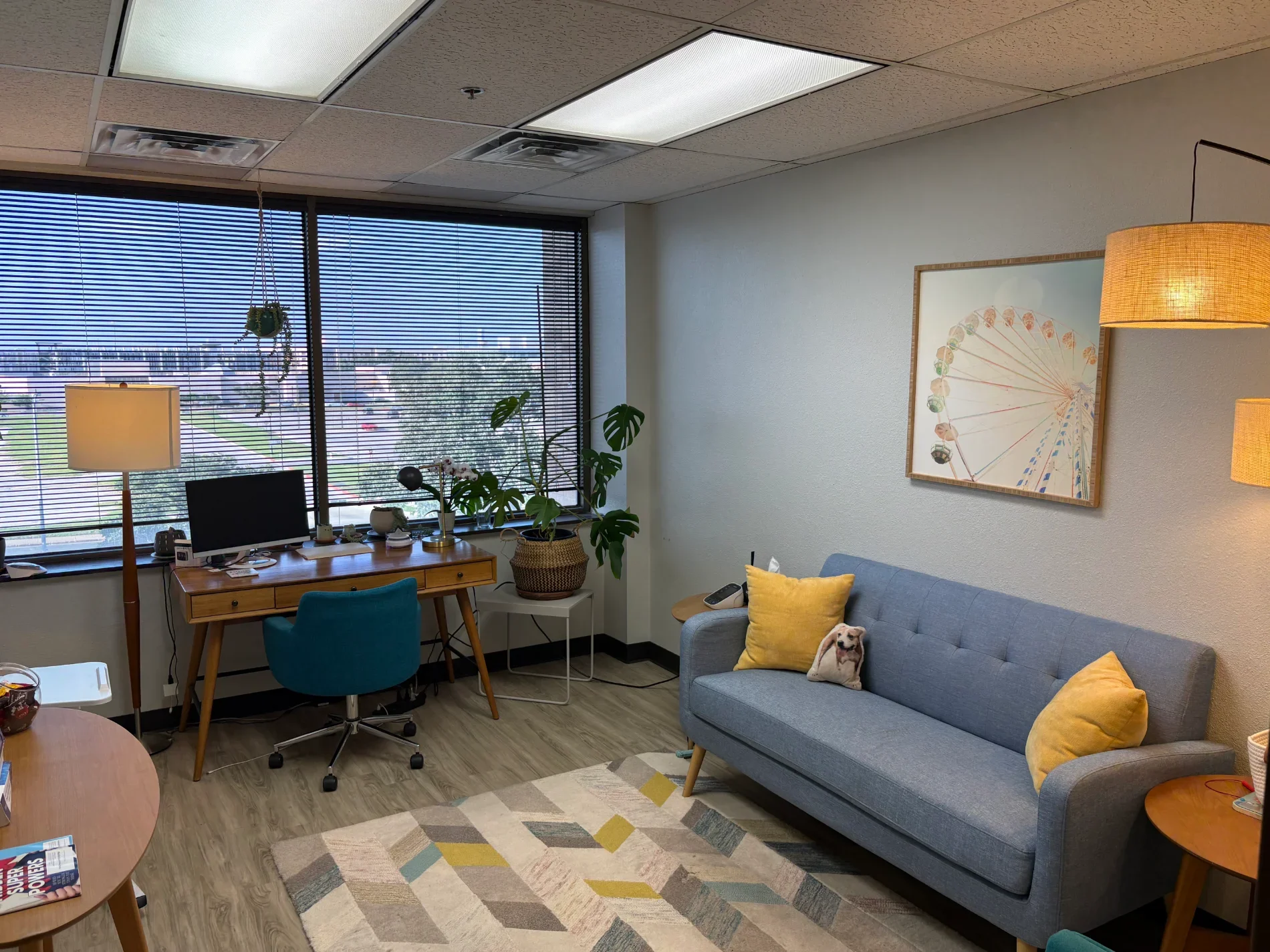 A room with large windows, plants, a grey sofa with yellow pillows, a wooden desk with a computer, a floor lamp, and wall art of a Ferris wheel.