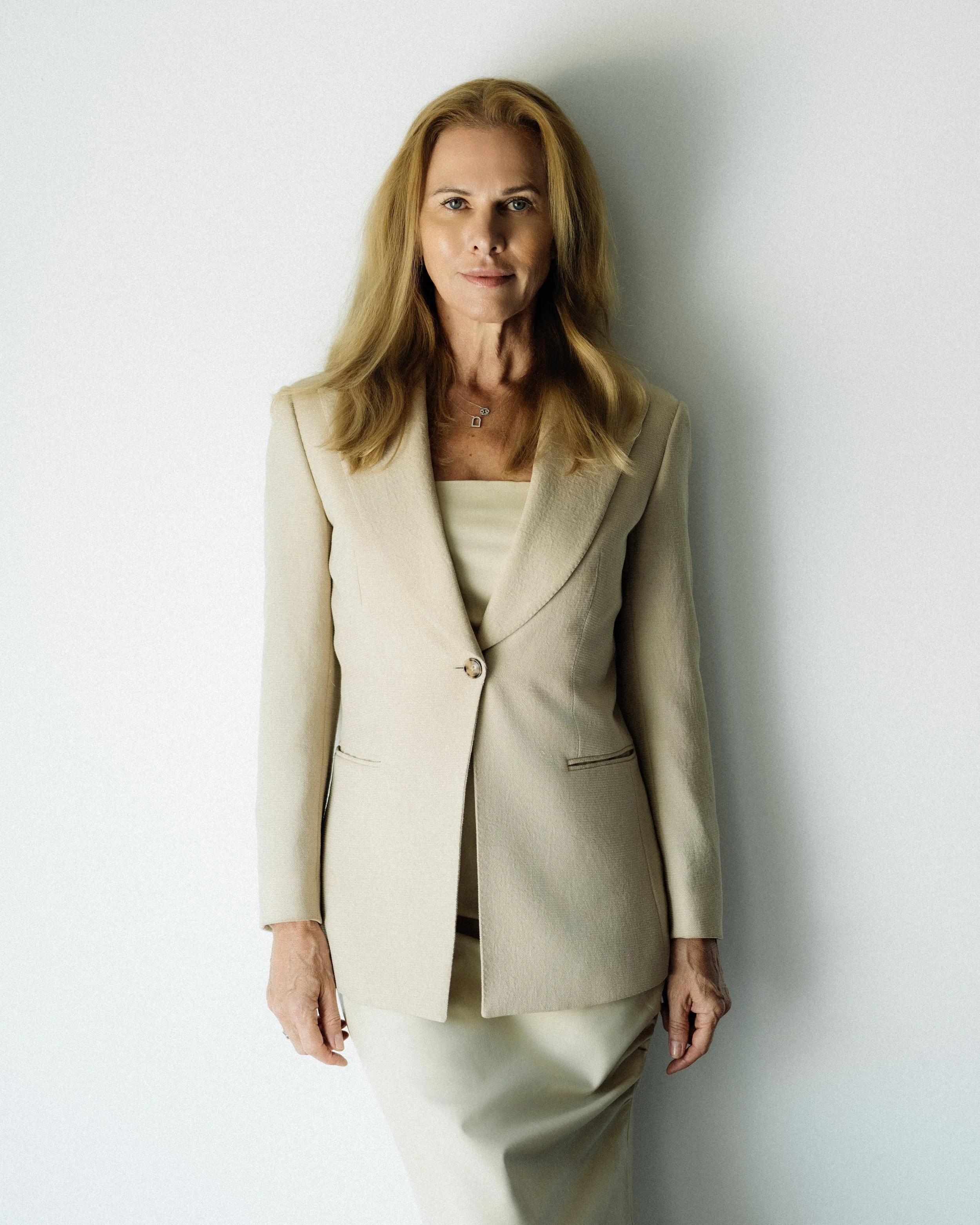 A woman with long blonde hair wearing a beige blazer and matching skirt, standing against a plain white wall.
