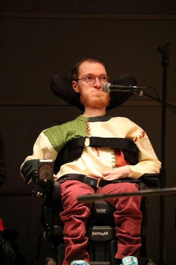 Stephen Verdile, a white man with red hair and a beard, sits in a motorized wheelchair in front of a microphone. He has round glasses, a patchwork sweater and red pants.