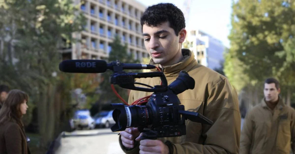 Adam Villani, a man with short black hair and a tan windbreaker, holds a cine camera while filming outside, focused on the shot.
