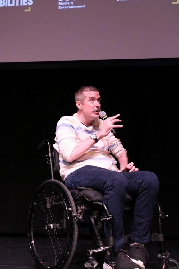 "Actor Michael Patrick Thornton, a white man with short, light-colored hair, a cream-colored sweater and navy pants sits in his manual wheelchair, speaking into a microphone on a stage.