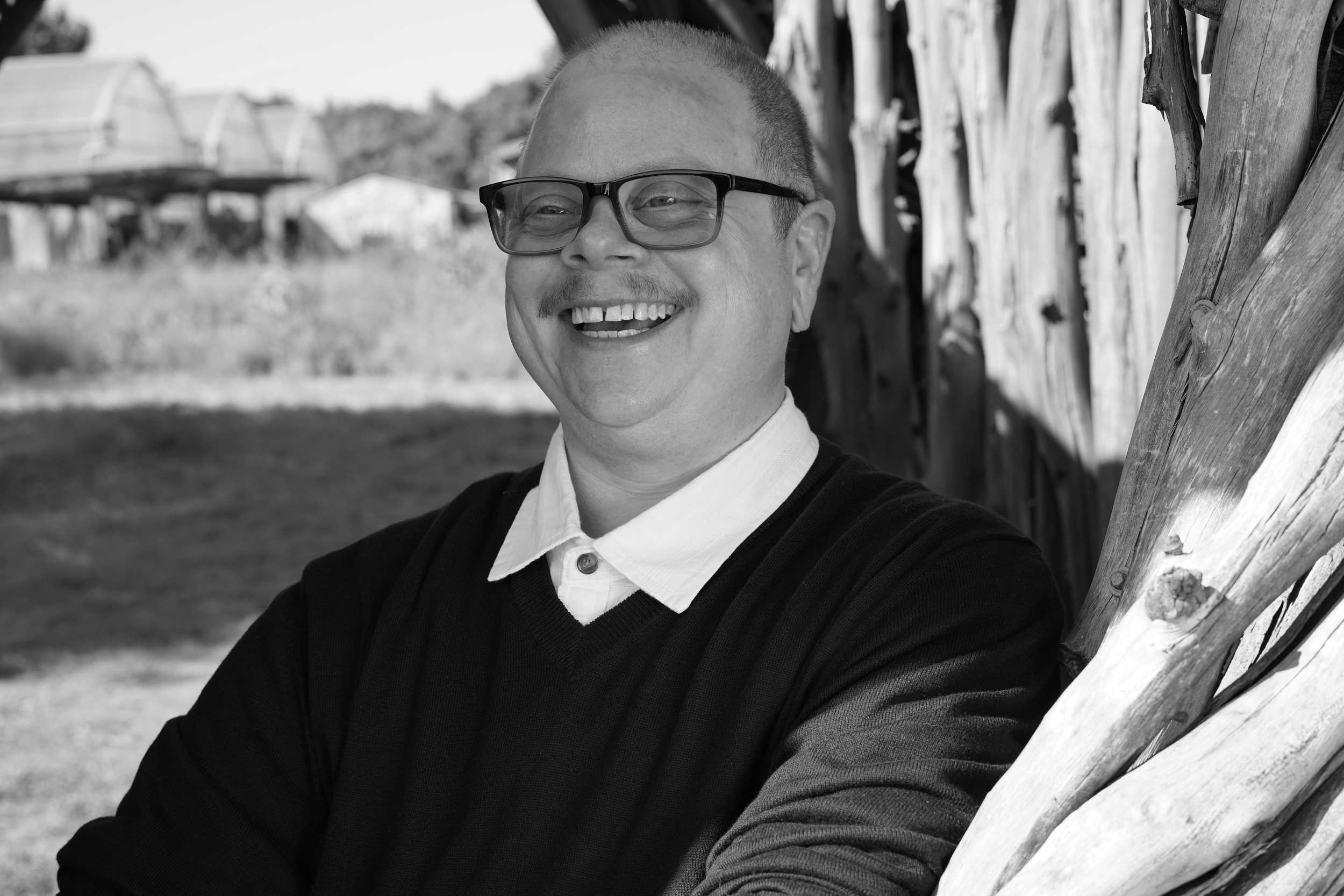 In black and white, Vega Darling is a non-binary person with a shaved head, glasses, a moustache and a vest over a collared shirt. He smiles widely.