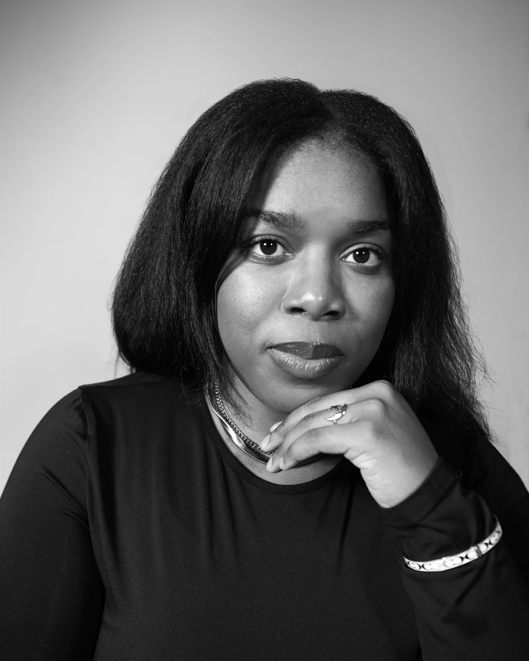 Black and white photo of brittany with her hand to chin looking directly act camera. She has black straight hair, a black long sleeve