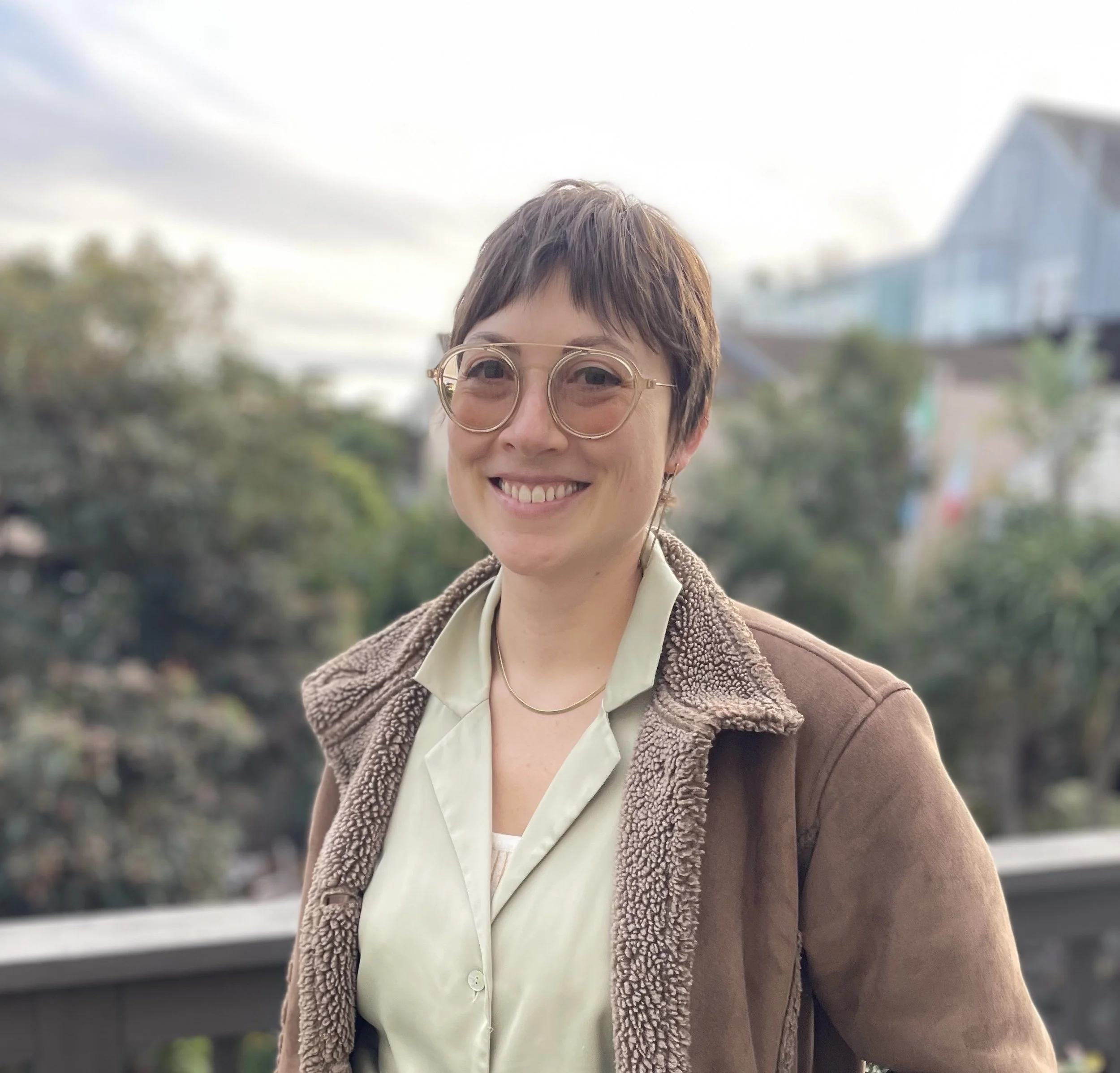 Maya Wise is a woman with large glasses, middle-parted bangs and and a casual button-up, covered by a zip-up hoodie. They stand outside, lush trees behind them.