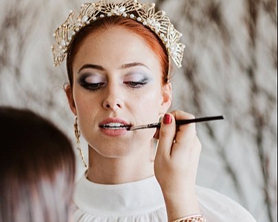 Makeup artist applying lip colour to a model with statement eye makeup and a decorative headpiece during a photo shoot..