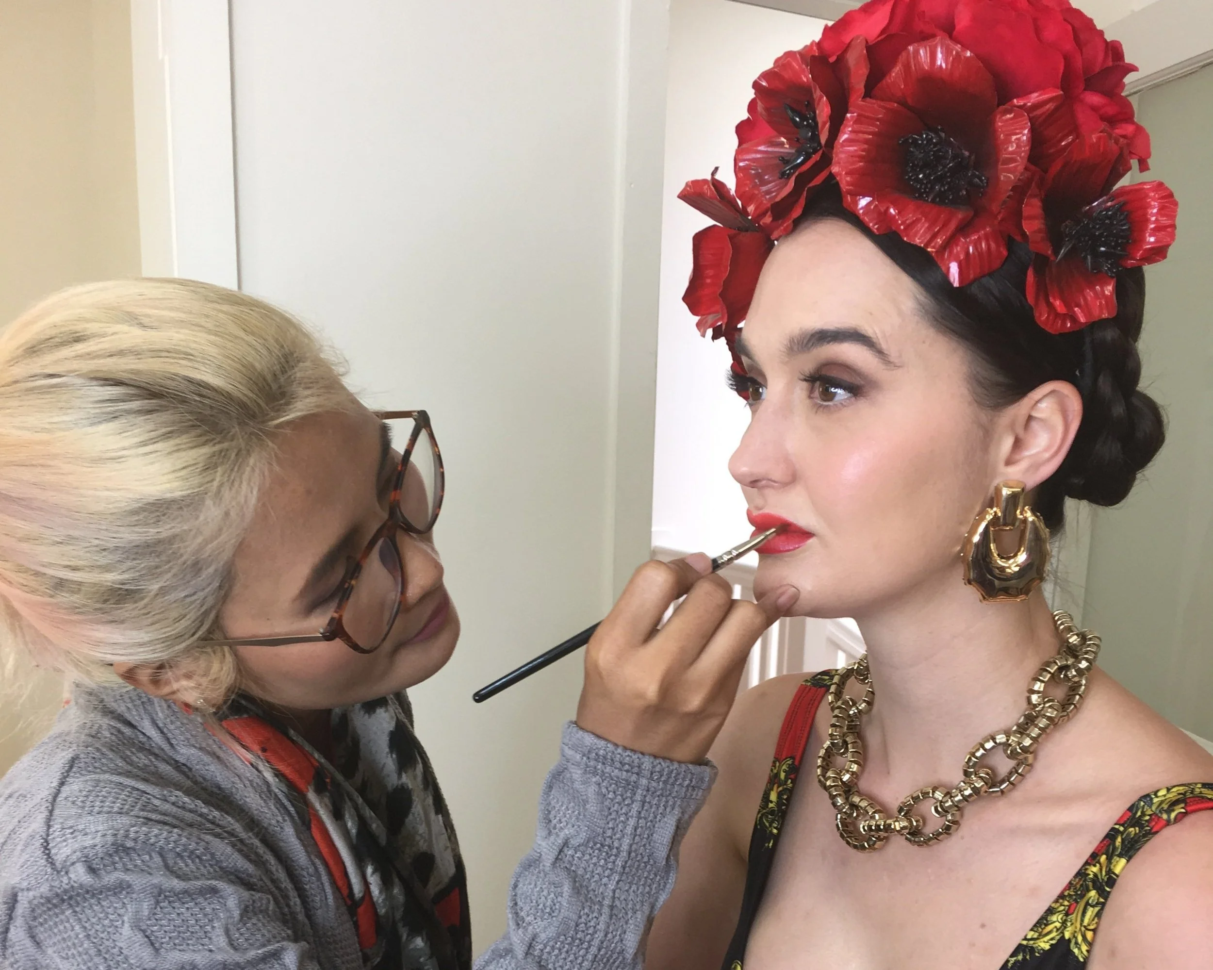 Makeup artist applying lipstick to a model wearing a bold floral headpiece and statement jewellery during a creative beauty session.