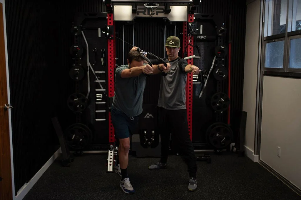 Two men training with resistance bands in a home gym, with weight plates and fitness equipment in the background.