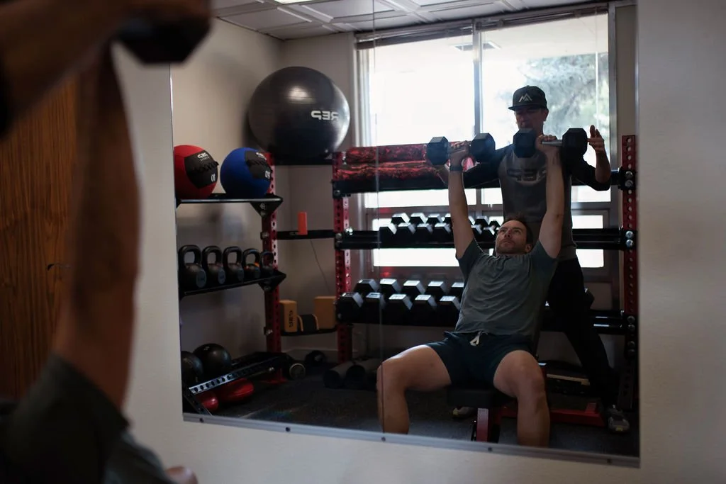 A client exercises with a dumbbell while sitting on a bench, guided by expert professional trainer Nick Gould in his private Durango gym. The gym has various weights, medicine balls, and fitness equipment. The scene is viewed through a mirror.