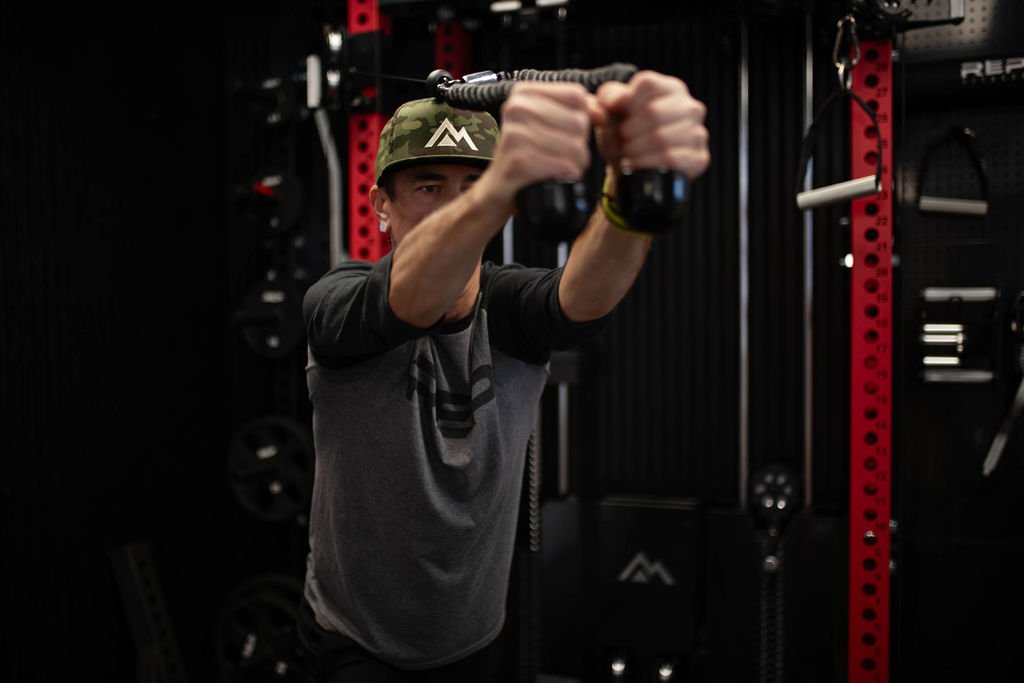 Nick Gould founder and owner of PurEnergy Performance lifting weights in a gym, holding a handle attachment with both hands, wearing a camouflage cap and a dark-colored t-shirt.