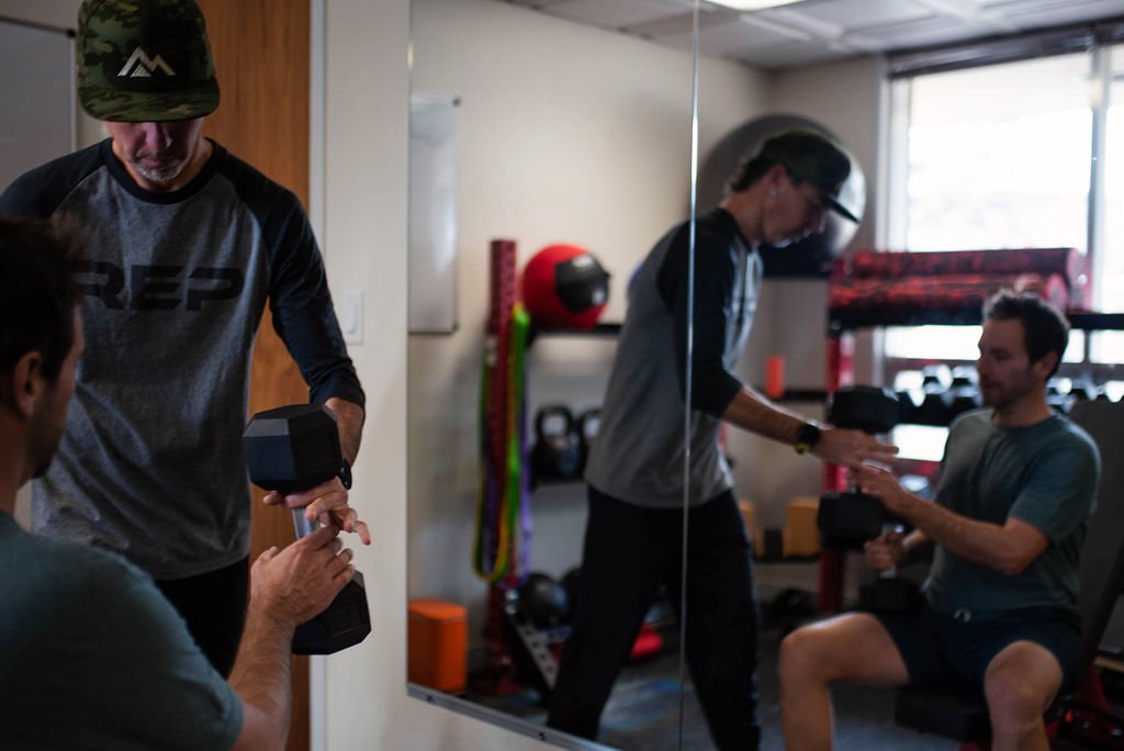 Nick Gould certified personal trainer and his client are training with dumbbells in the Durango gym, with one sitting and the other standing.