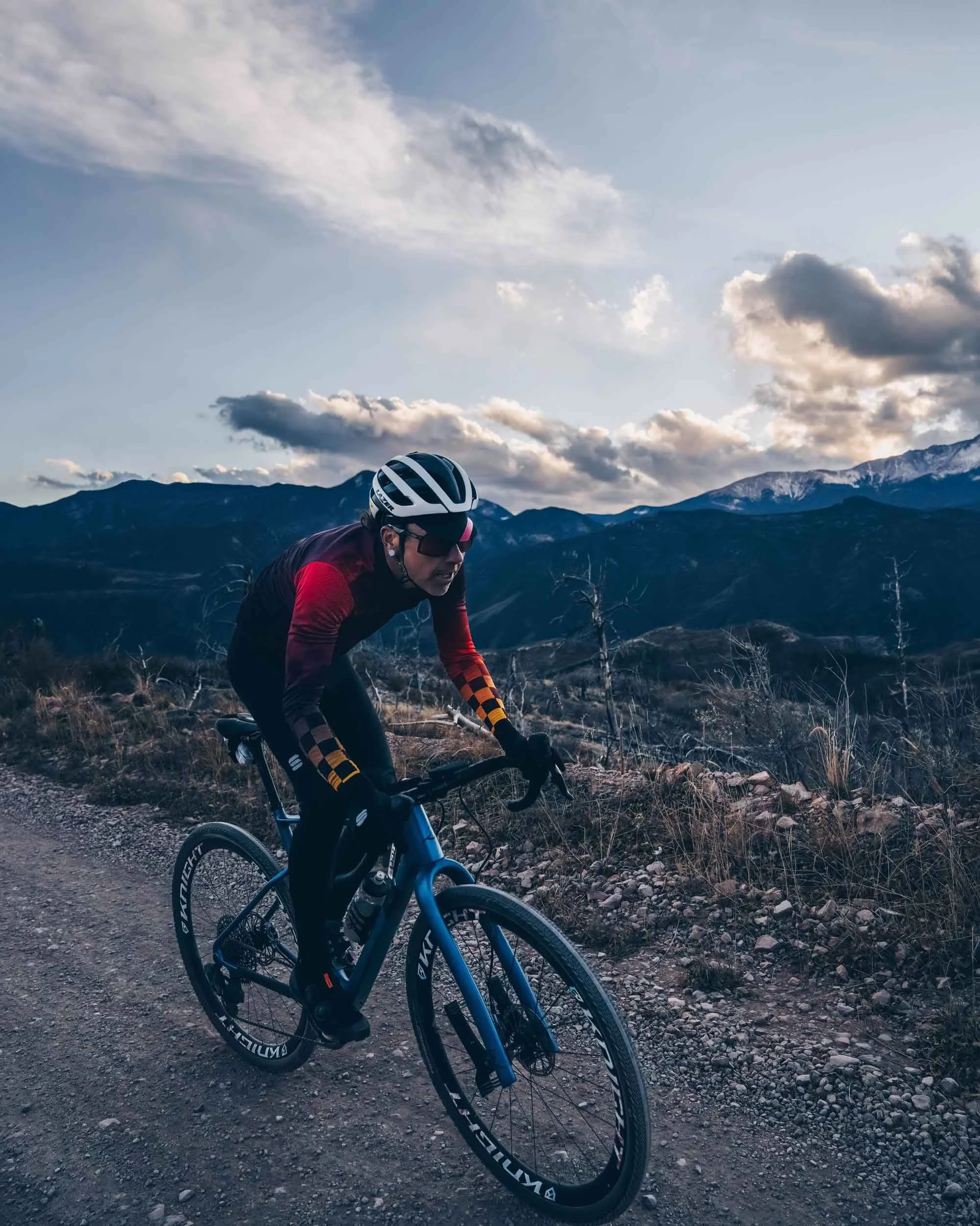 Nick Gould professional cyclist riding on a mountain trail during daylight, with mountains and cloudy sky in the background.